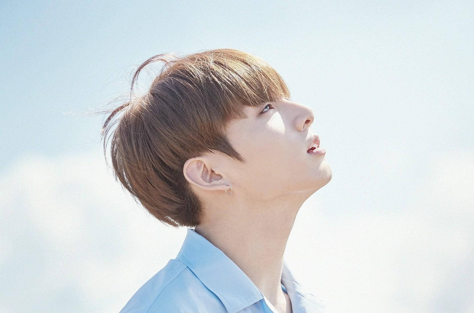 Jungkook 桌布 壁紙 — 一位棕色頭髮、身穿淺藍色襯衫的年輕男子，在柔和雲朵的明亮天空下向上凝視。