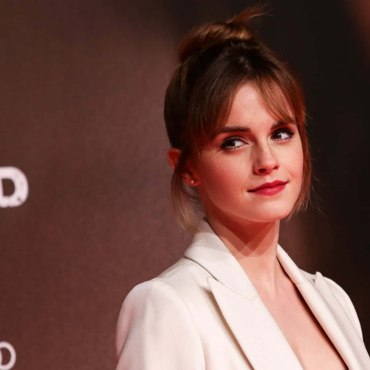 Emma Watson 桌布 壁紙 — 一位棕色頭髮凌亂盤成髮髻的女性，身穿白色西裝外套，在深色背景下望向一旁。