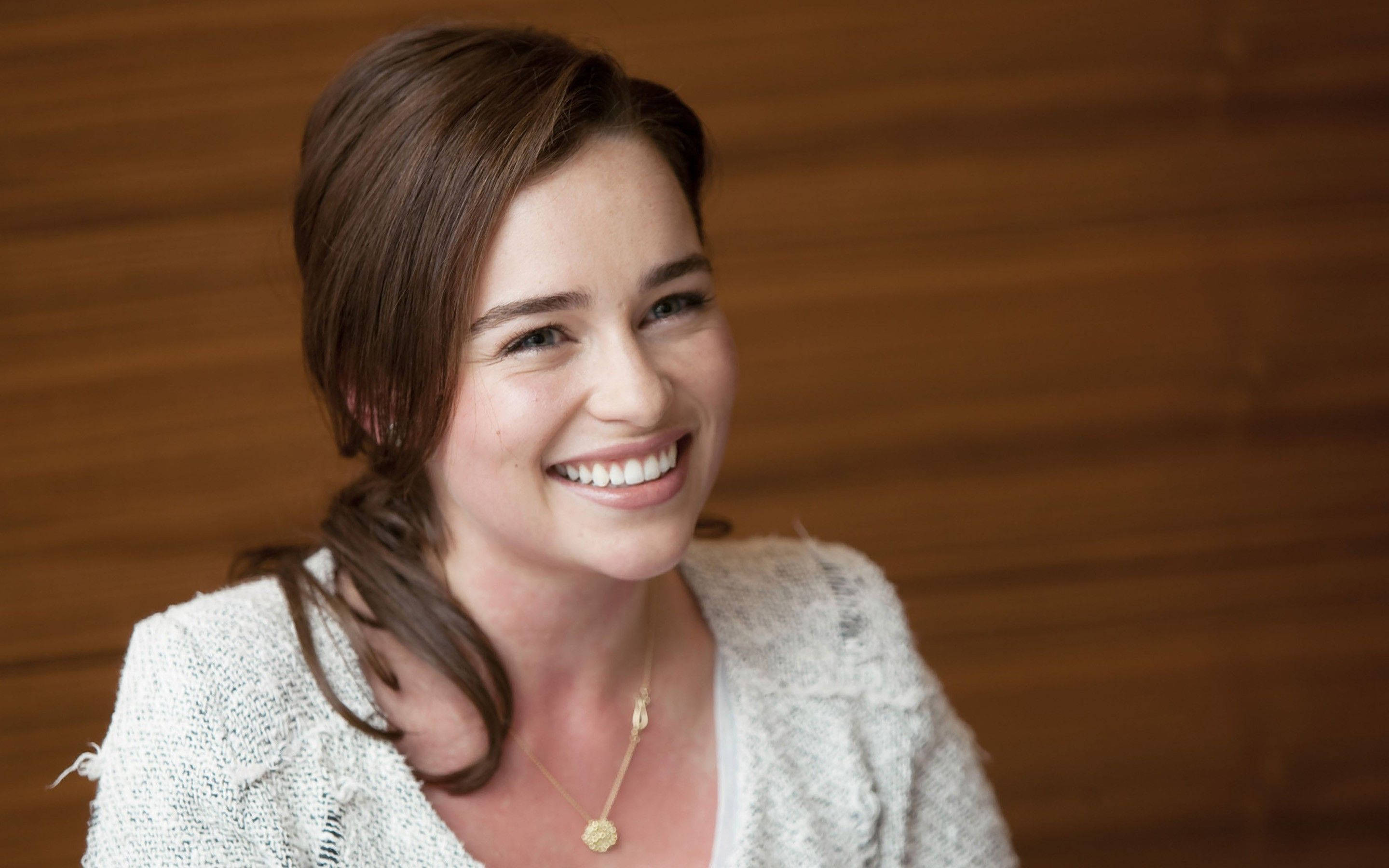 Emilia 桌布 壁紙 — 一位棕色頭髮的女性在木質背景前溫暖微笑，穿著一件有紋理的白色開襟衫。