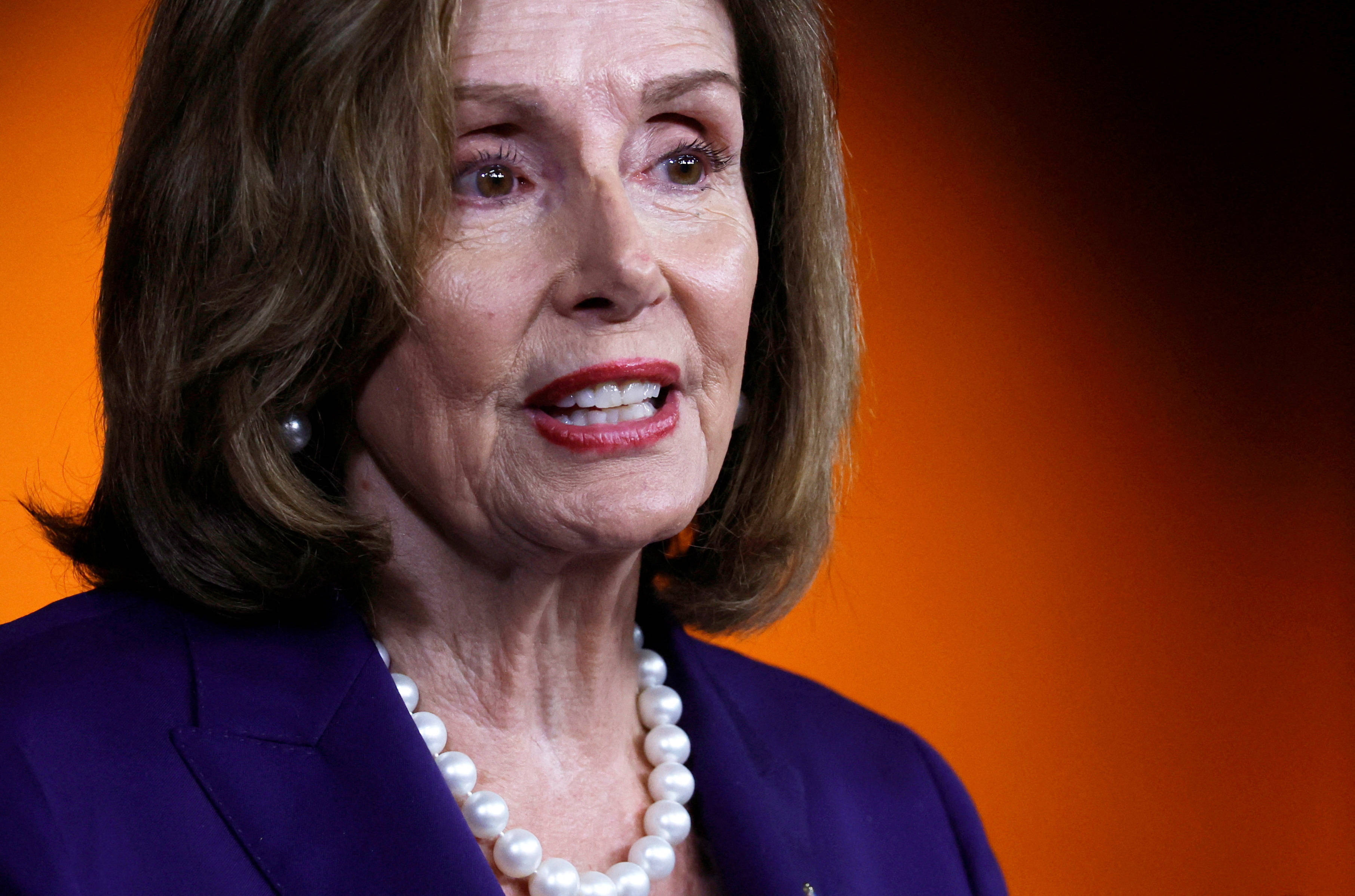 Nancy Pelosi 桌布 壁紙 — 一位棕色頭髮的女性穿著紫色西裝和珍珠項鍊，在溫暖的橙色背景下的特寫肖像。