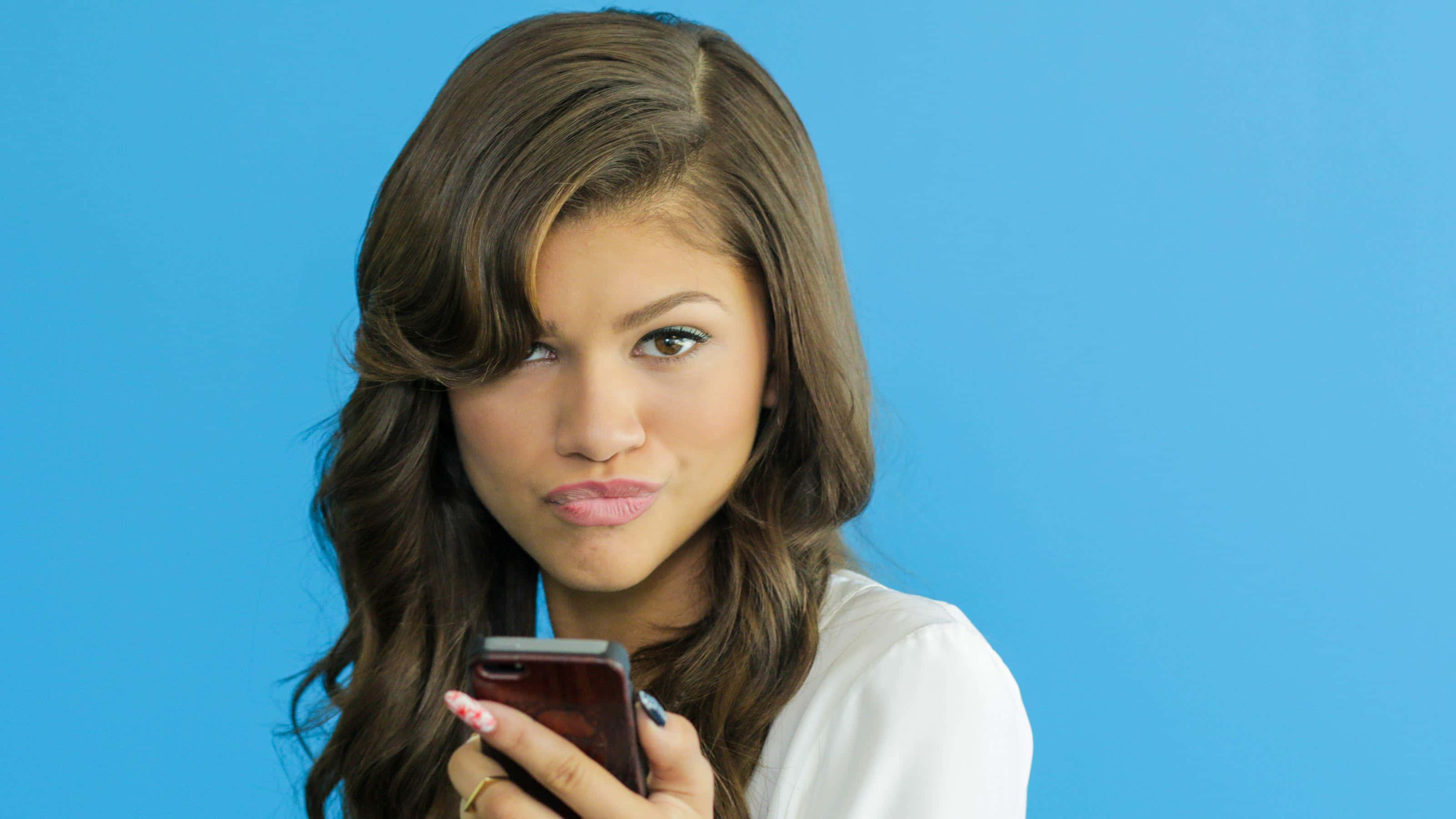 Zendaya 桌布 壁紙 — 一位棕色頭髮的年輕女性在純藍色背景前拿著手機，直接看著鏡頭，表情俏皮。