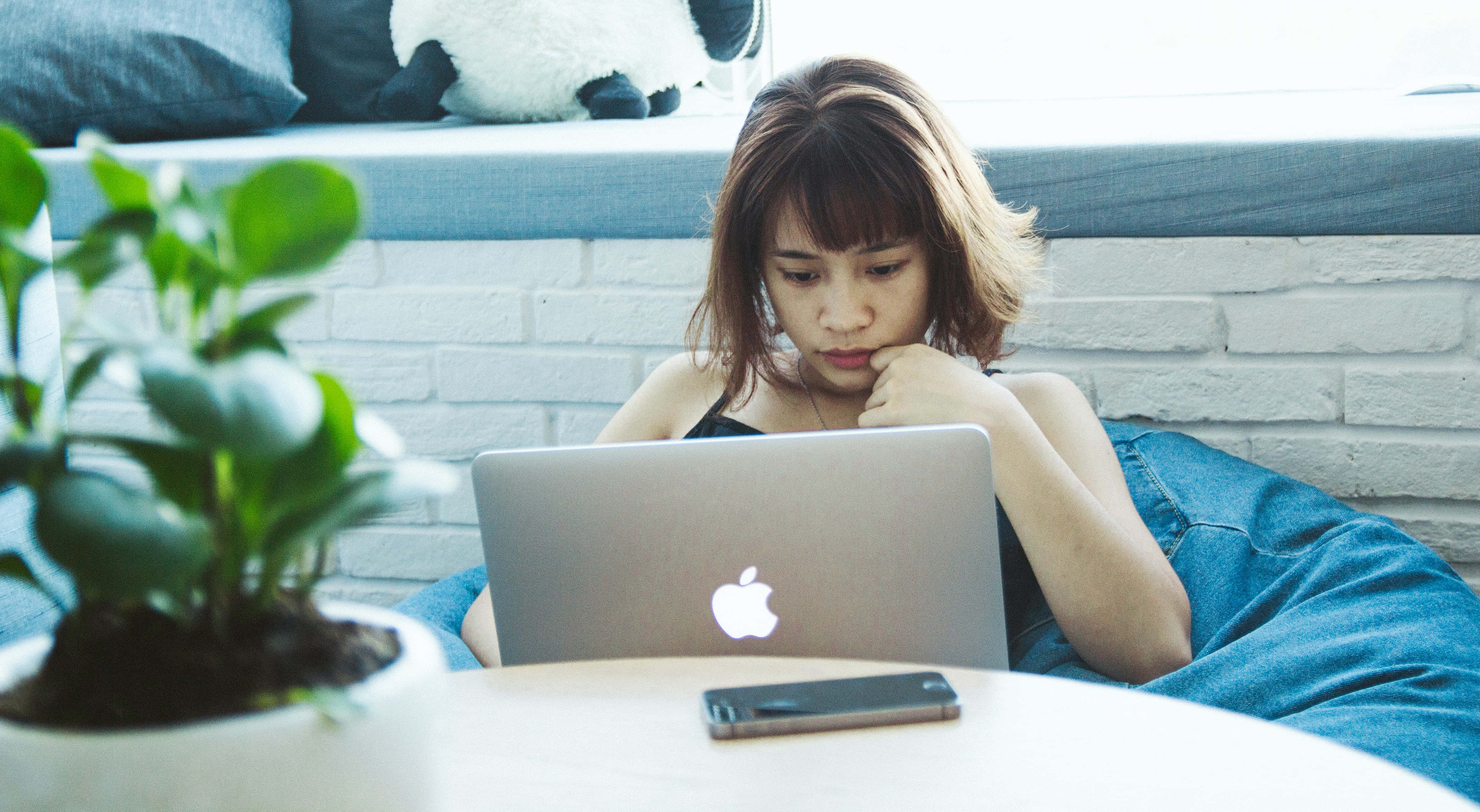 美學 MacBook 壁紙 桌布 壁紙 — 一位棕色頭髮的年輕女性坐在藍色豆袋椅上，專注於她的筆記型電腦，房間明亮，附近有一盆盆栽植物。
