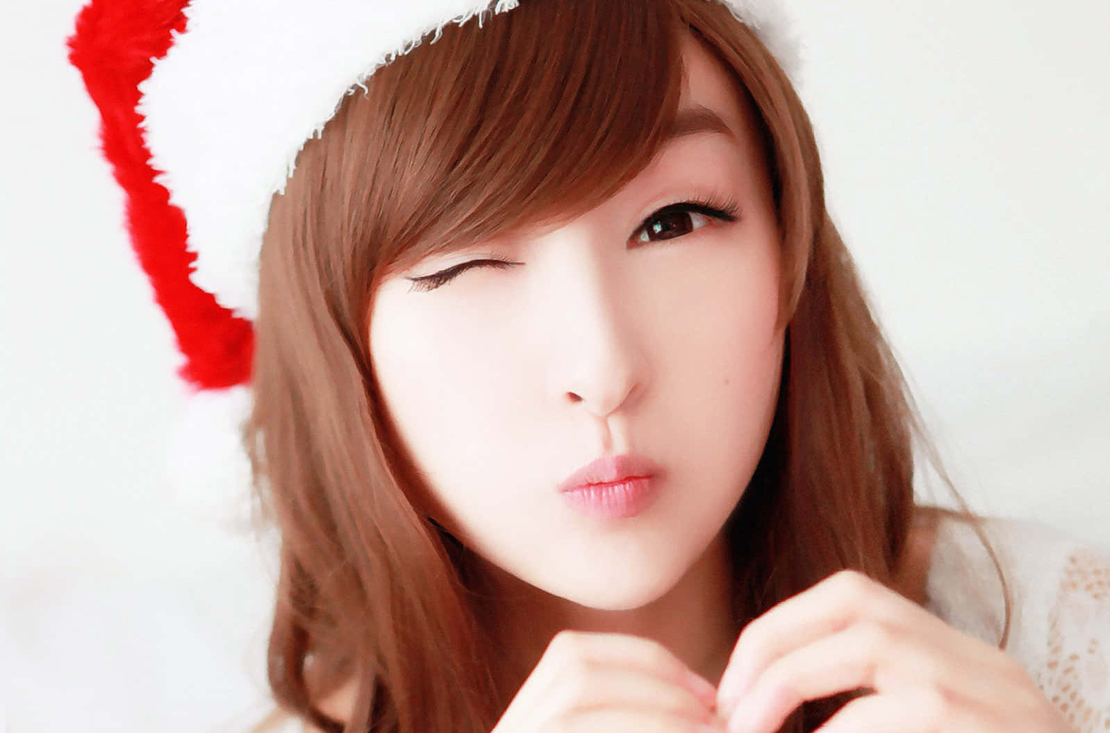 Santa 桌布 壁紙 — 一位棕色頭髮的年輕女性戴著紅白相間的聖誕帽，在明亮的白色背景前俏皮地眨眼。