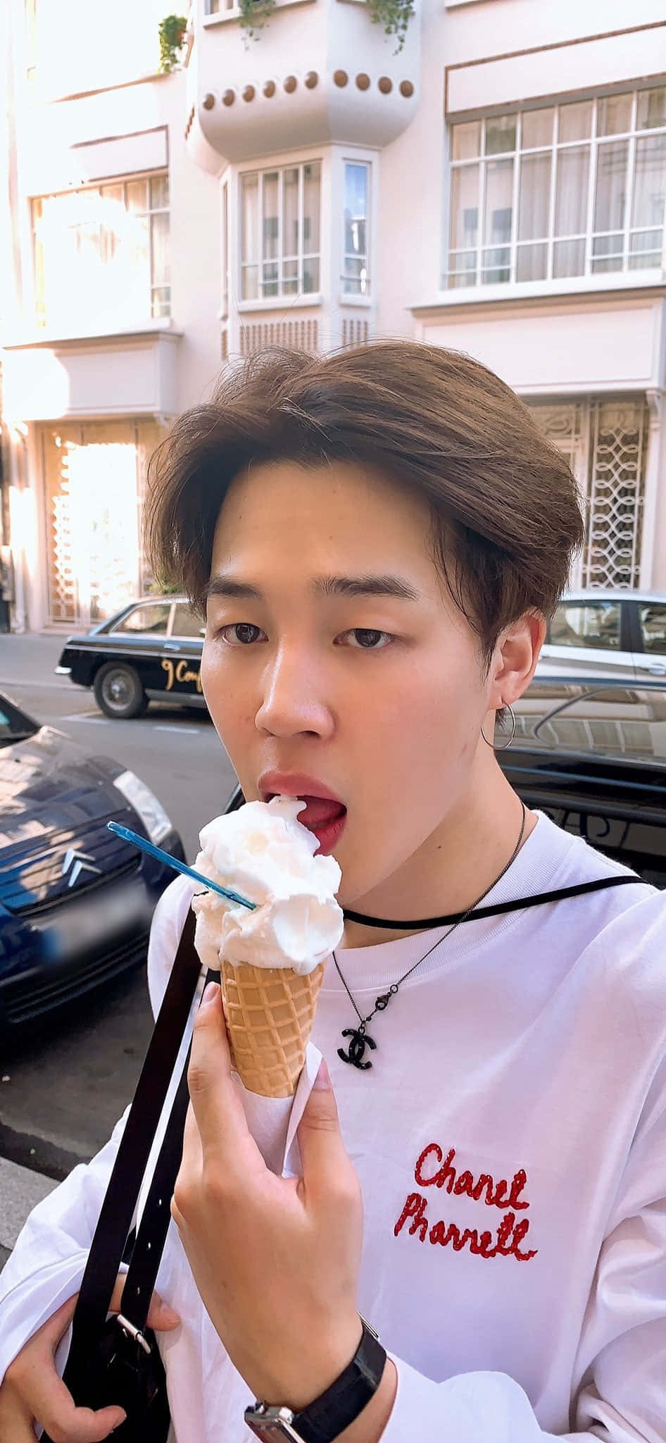 Jimin Aesthetic 桌布 壁紙 — 一位棕色頭髮的年輕男子在陽光明媚的城市街道上享用香草冰淇淋甜筒，身後有復古汽車和粉彩建築。