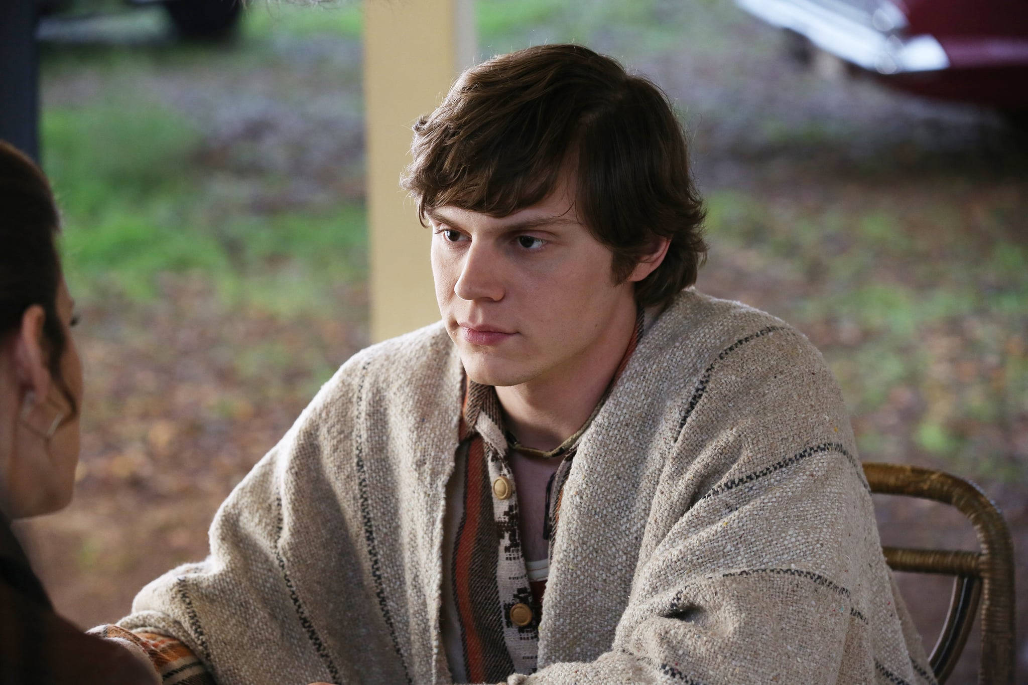 Tate Langdon 桌布 壁紙 — 一位棕色頭髮的年輕男子坐在藤椅上，穿著質感米色開襟衫和圖案襯衫，在柔和光線的戶外環境中專注地看著鏡頭外的某人。