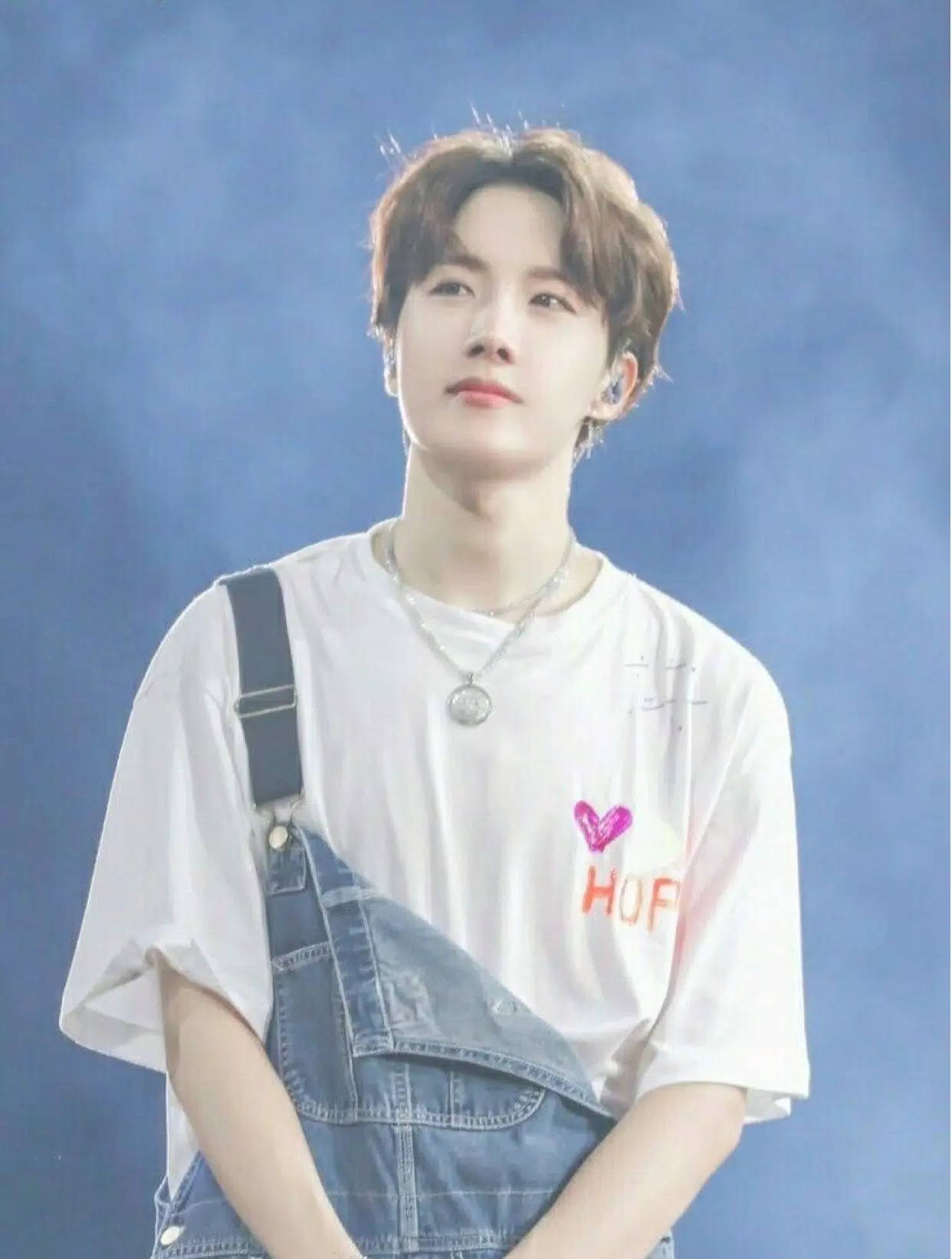 jhope 桌布 壁紙 — 一位棕色頭髮的年輕男子穿著白色T恤，上面有粉紅色心形和橙色文字，外面套著藍色牛仔吊帶褲，背景是模糊的藍色。