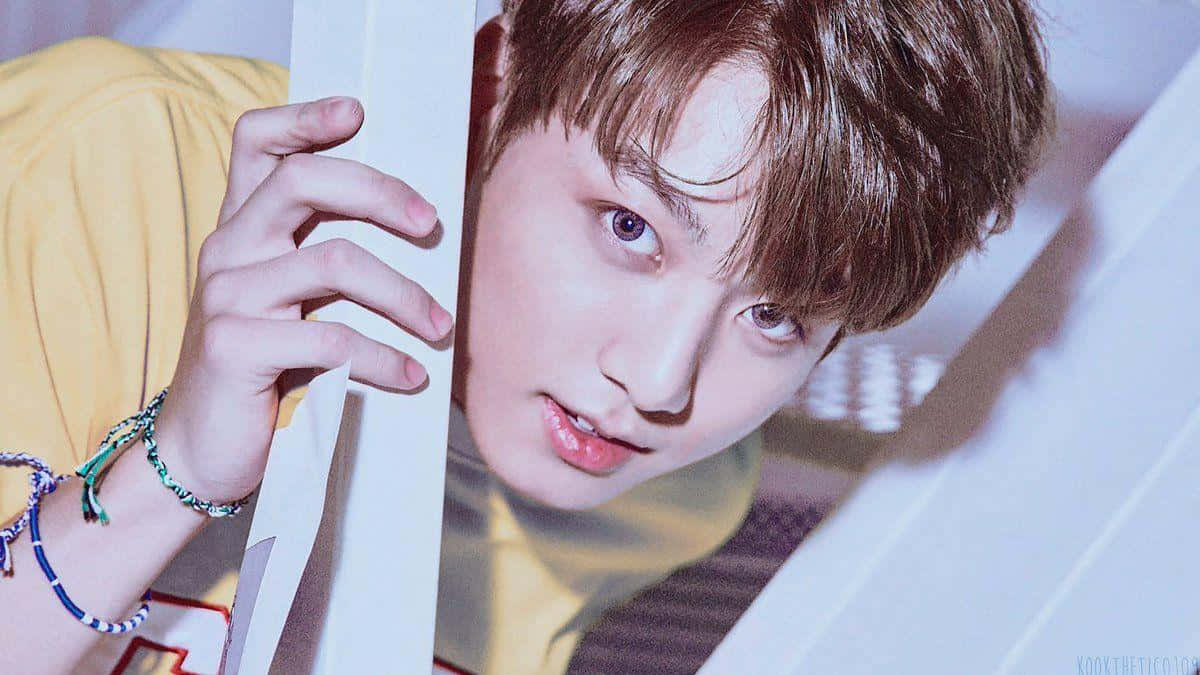Jungkook 桌布 壁紙 — 一位棕色頭髮的年輕男子穿著黃色襯衫和彩色手鐲，透過白色板條屏障窺視。