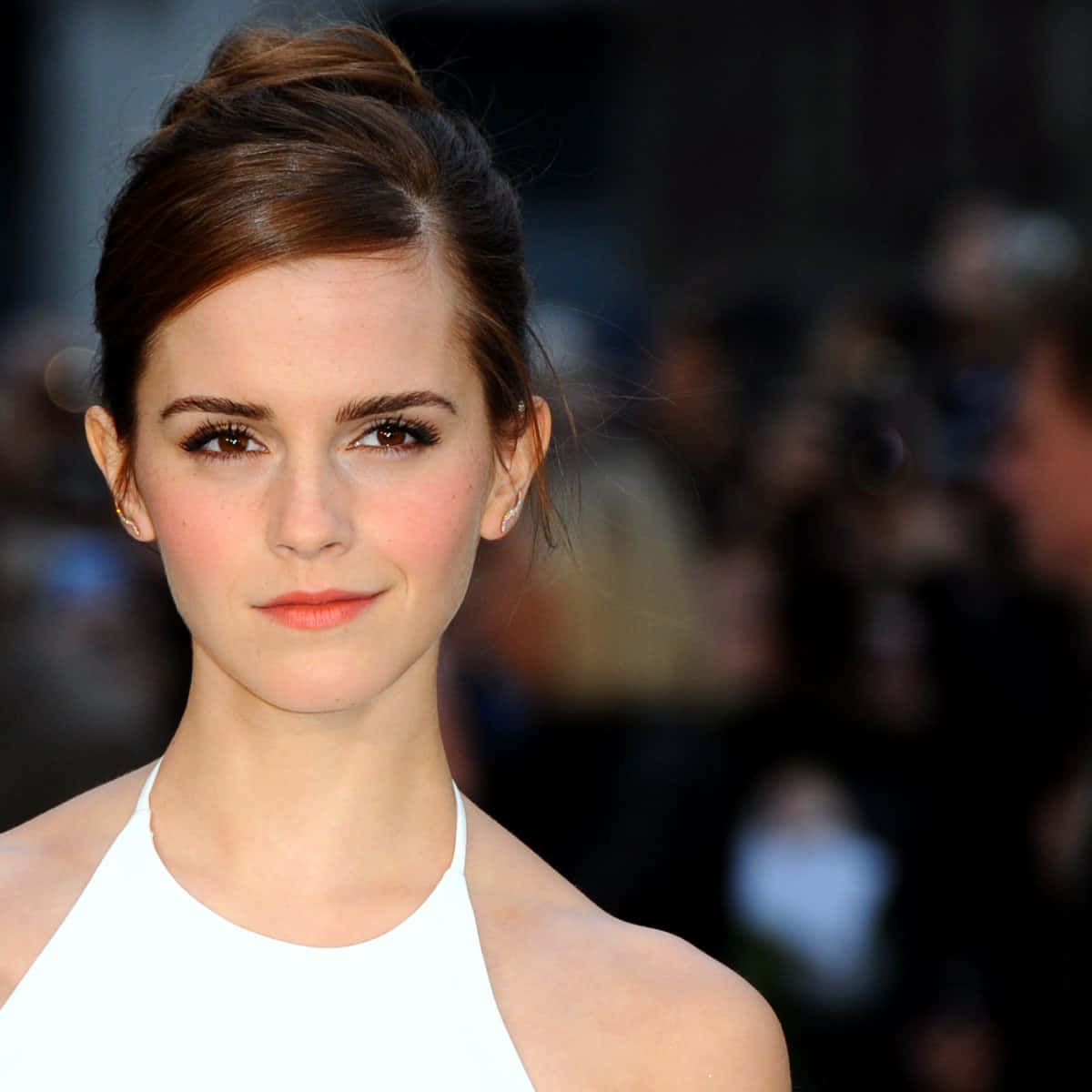 Emma Watson 桌布 壁紙 — 一位棕色頭髮盤起的女性身穿白色掛頸上衣，背景是模糊的人群的特寫肖像。