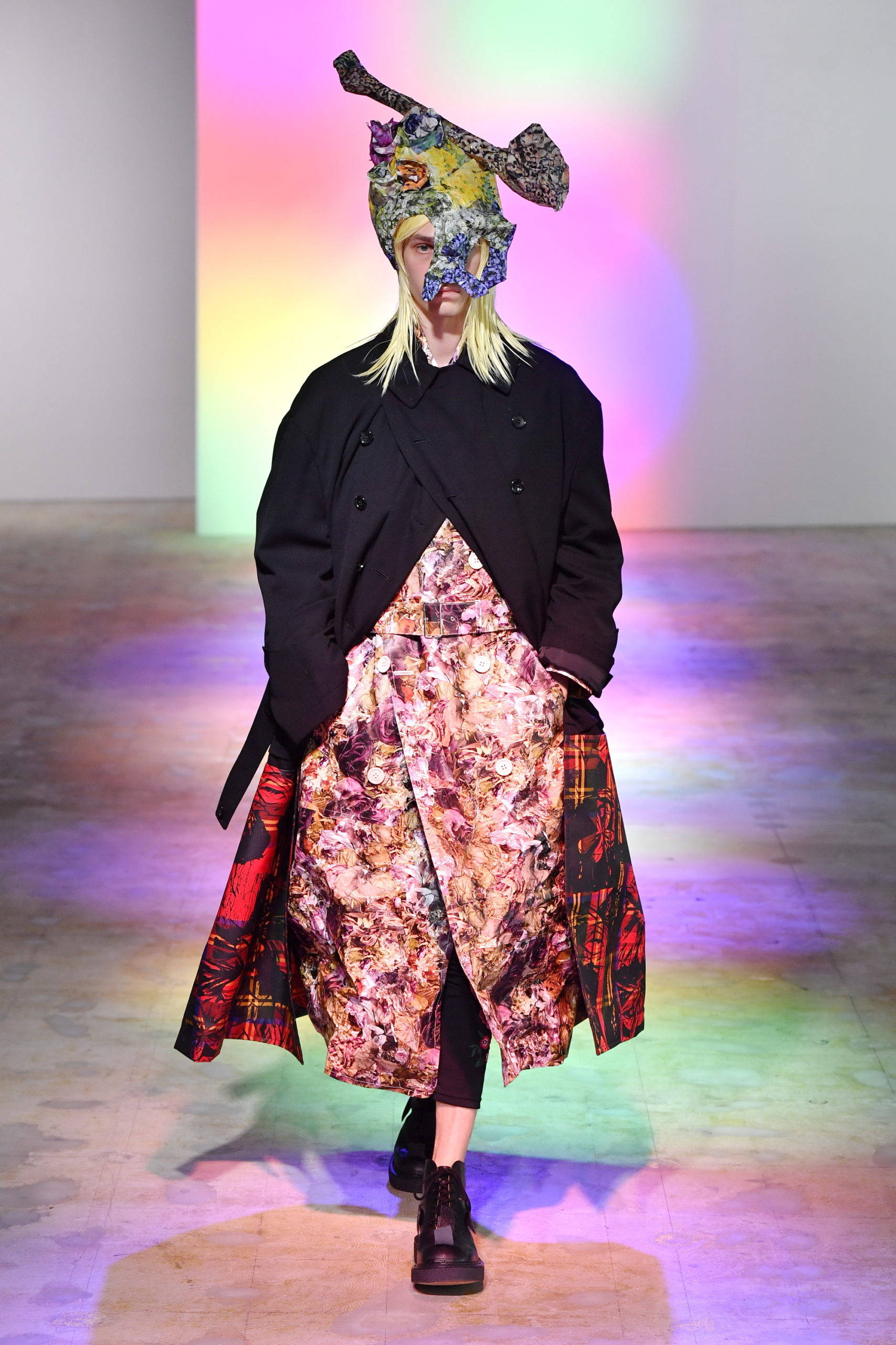 時尚 桌布 壁紙 — 一位模特兒在伸展台上行走，穿著黑色外套搭配花卉圖案裙子，並在充滿活力的舞台燈光下戴著色彩繽紛的圖案口罩。