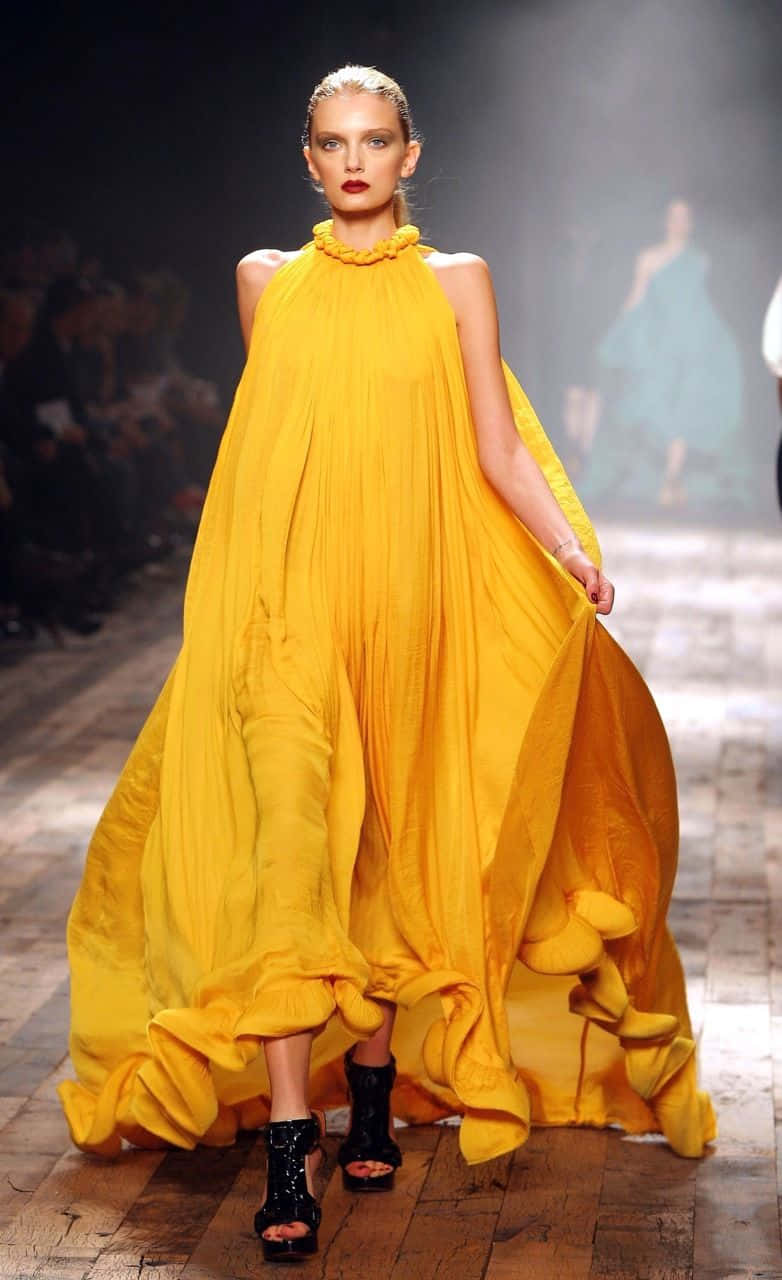 Yellow Fashion 桌布 壁紙 — 一位模特兒在戲劇性的舞台燈光下，自信地穿著一件寬大飄逸的黃色長裙走過伸展台。