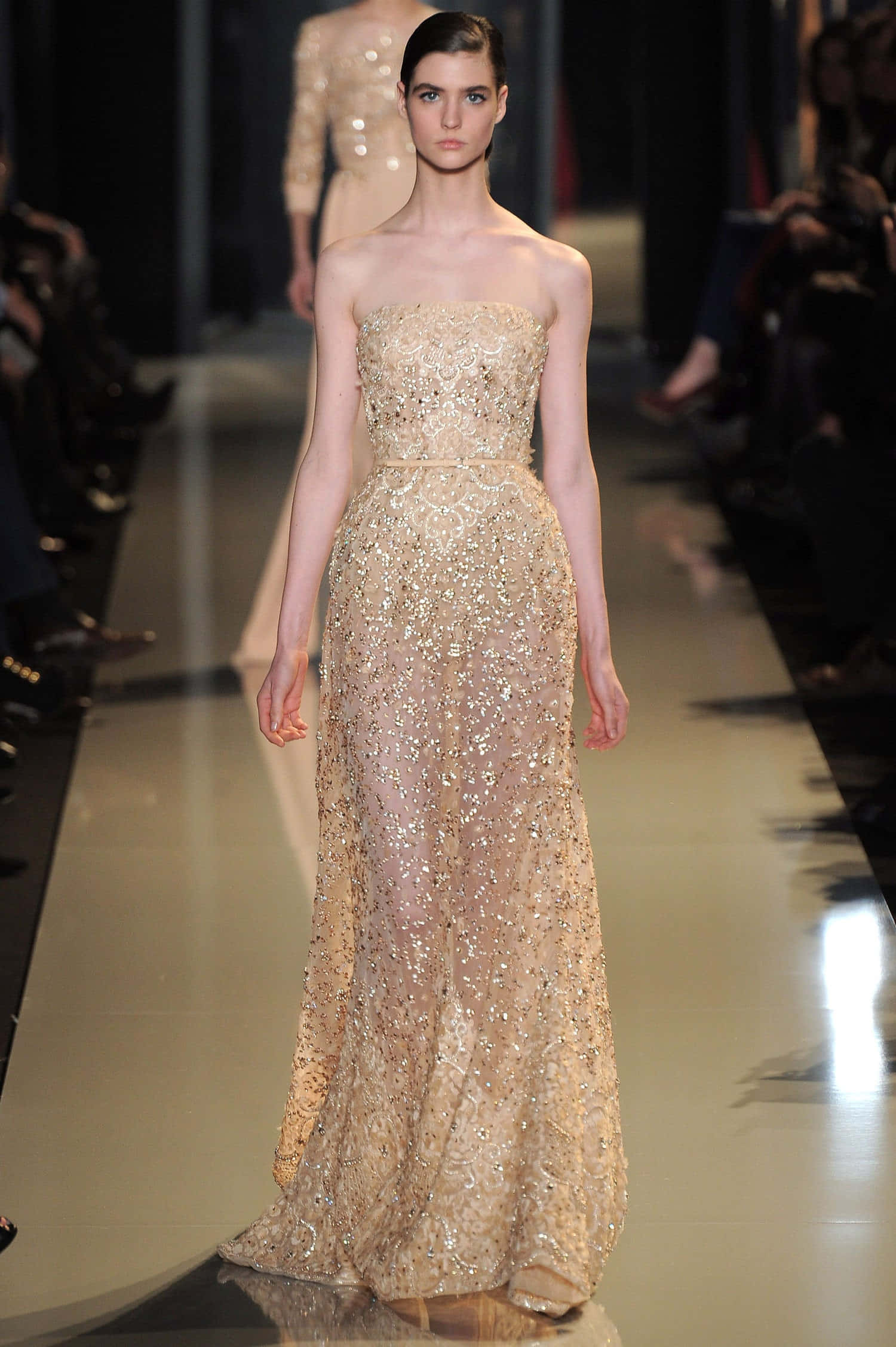 Beautiful Fashion 桌布 壁紙 — 一位模特兒在明亮的舞台燈光下，穿著優雅的金色亮片無肩帶長裙，自信地走在伸展台上。