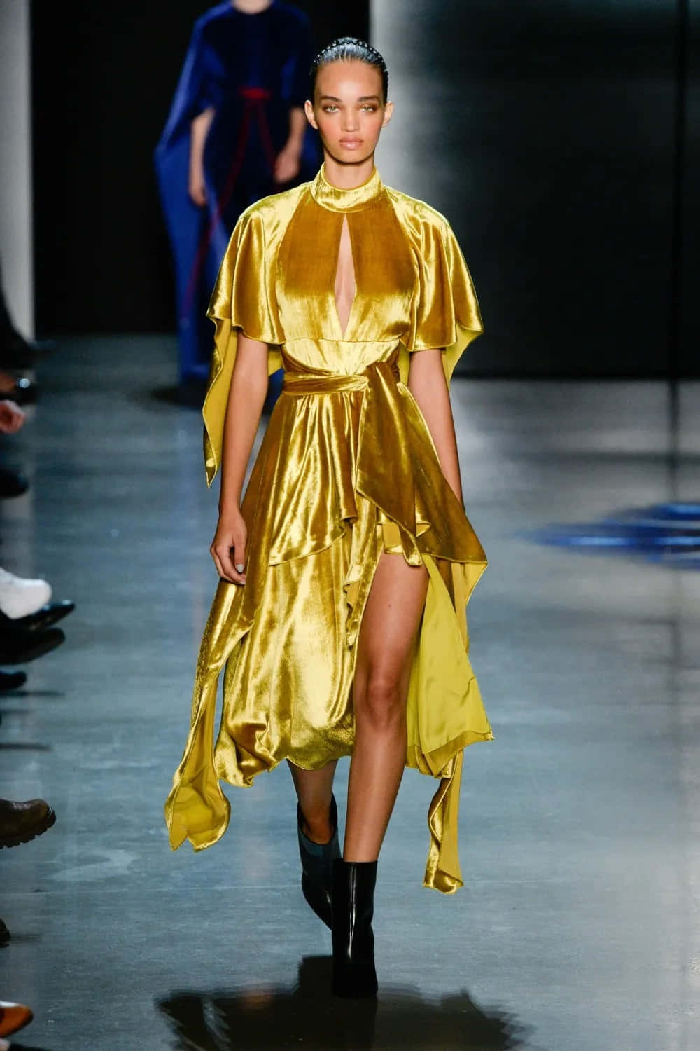 Yellow Fashion 桌布 壁紙 — 一位模特兒在明亮的舞台燈光下，自信地穿著閃閃發光的金色緞面連衣裙，帶有高開衩和斗篷袖，走在伸展台上。