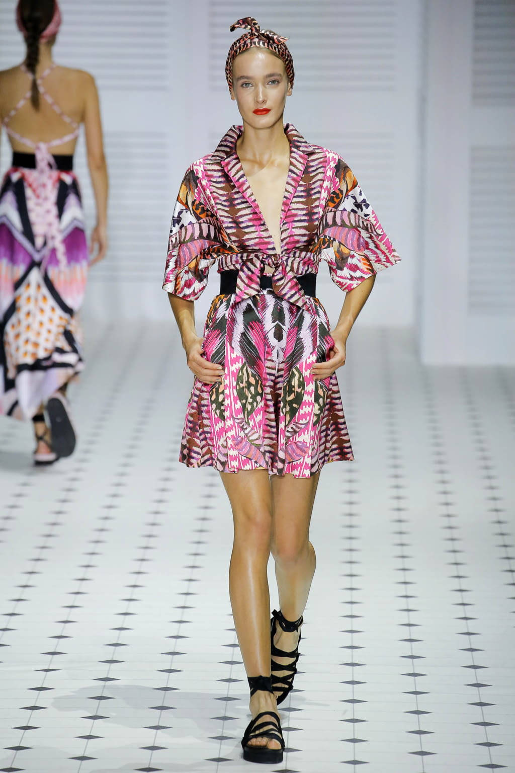 Runway 桌布 壁紙 — 一位模特兒在白色瓷磚跑道上穿著充滿活力的粉紅色和棕色圖案和服連衣裙搭配黑色高跟鞋走秀。