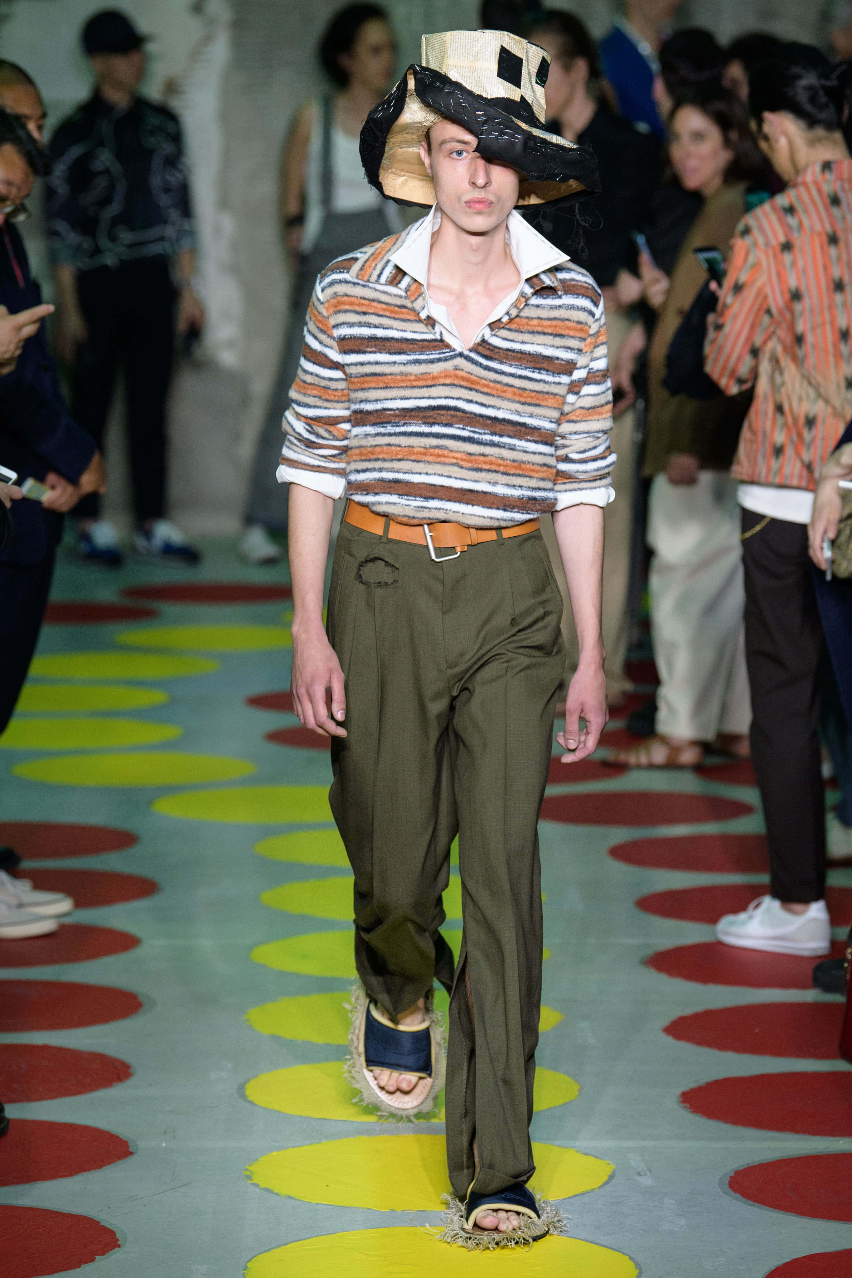 Runway 桌布 壁紙 — 一位模特兒在色彩繽紛的圓形圖案伸展台上行走，身穿條紋襯衫、橄欖色褲子和一頂大帽子。