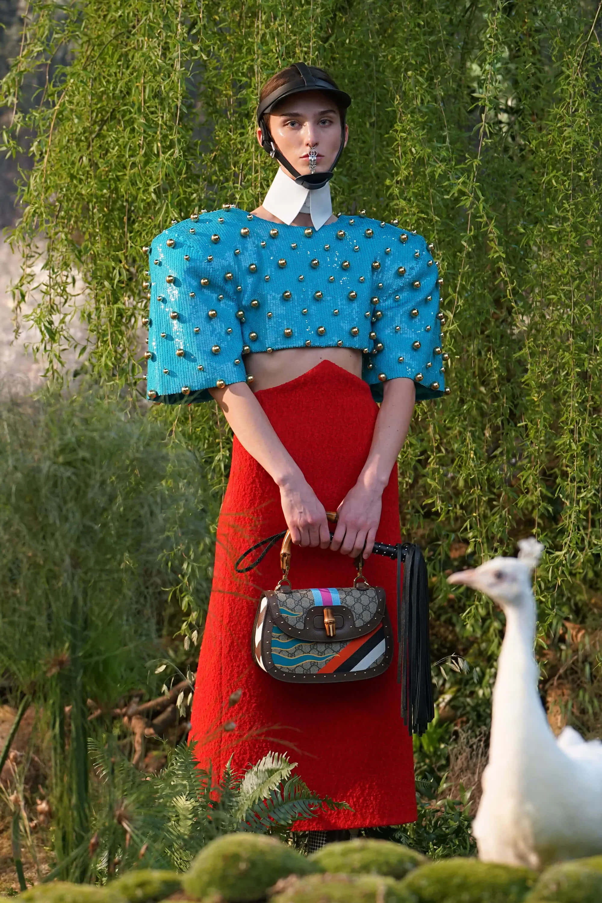 Gucci 桌布 壁紙 — 一位模特兒穿著大膽的藍色鉚釘露臍上衣和紅色裙子，站在茂密的綠植中，手持圖案手袋，附近有一隻白色孔雀。