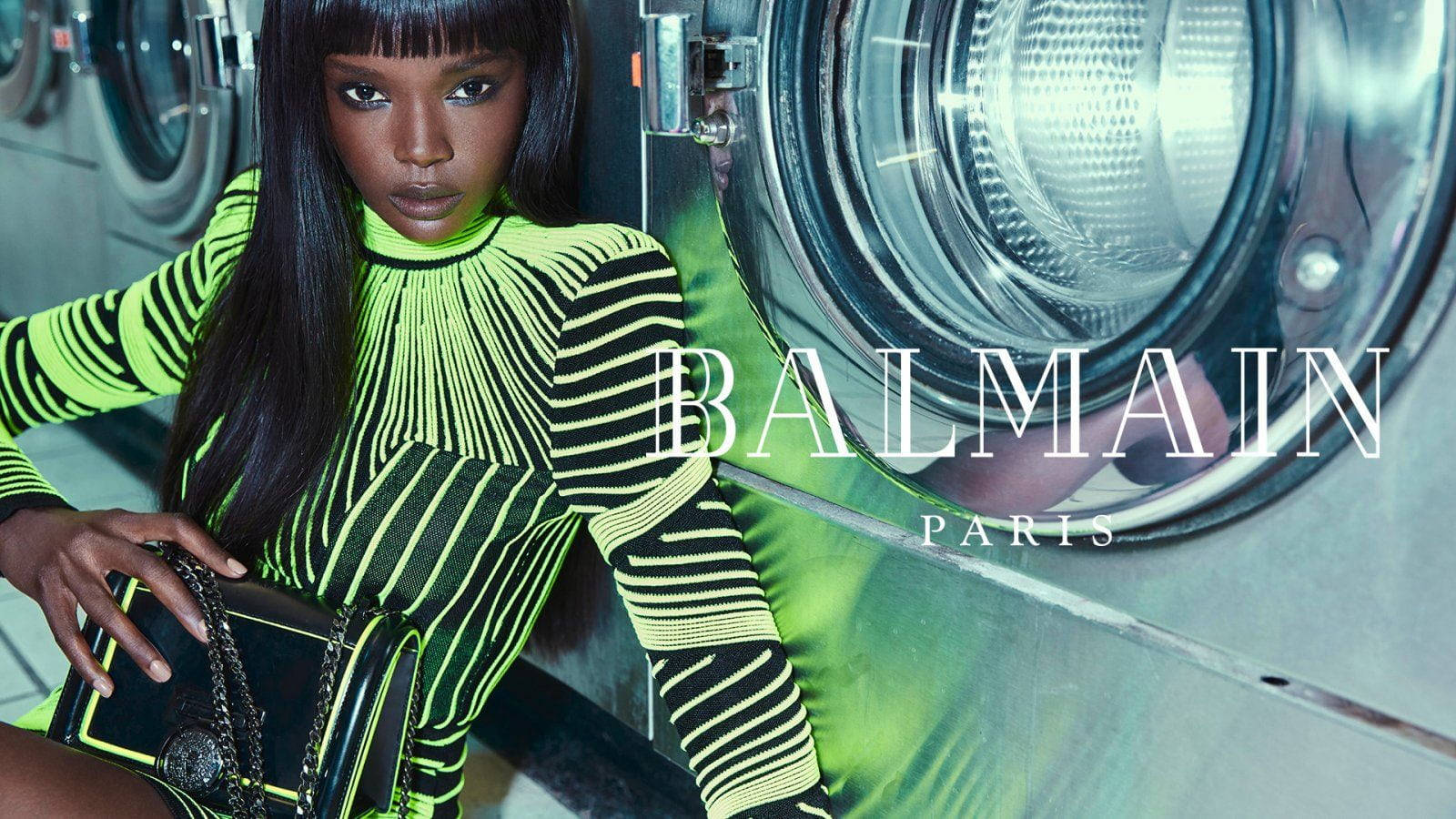 桌布 壁紙 — 一位模特兒穿著螢光綠色條紋服裝，在 Balmain Paris 時尚廣告中自信地在工業洗衣機旁擺姿勢。
