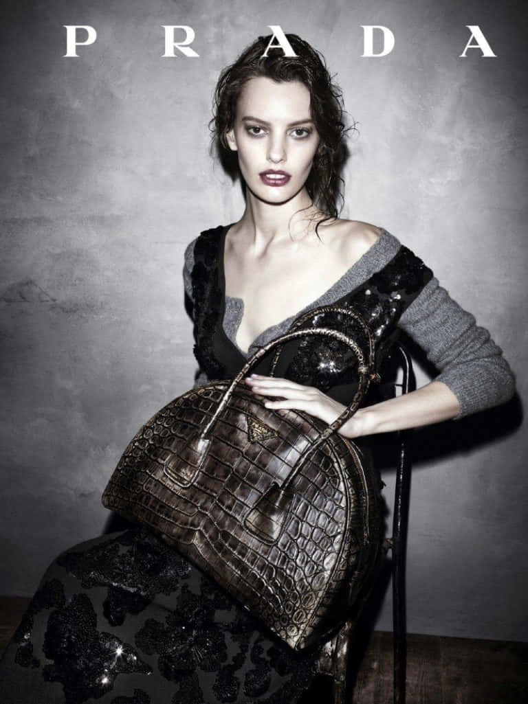 Prada 桌布 壁紙 — 一位模特兒穿著質感豐富的黑灰色服裝，在柔和的灰色工作室背景前拿著鱷魚皮Prada手袋。