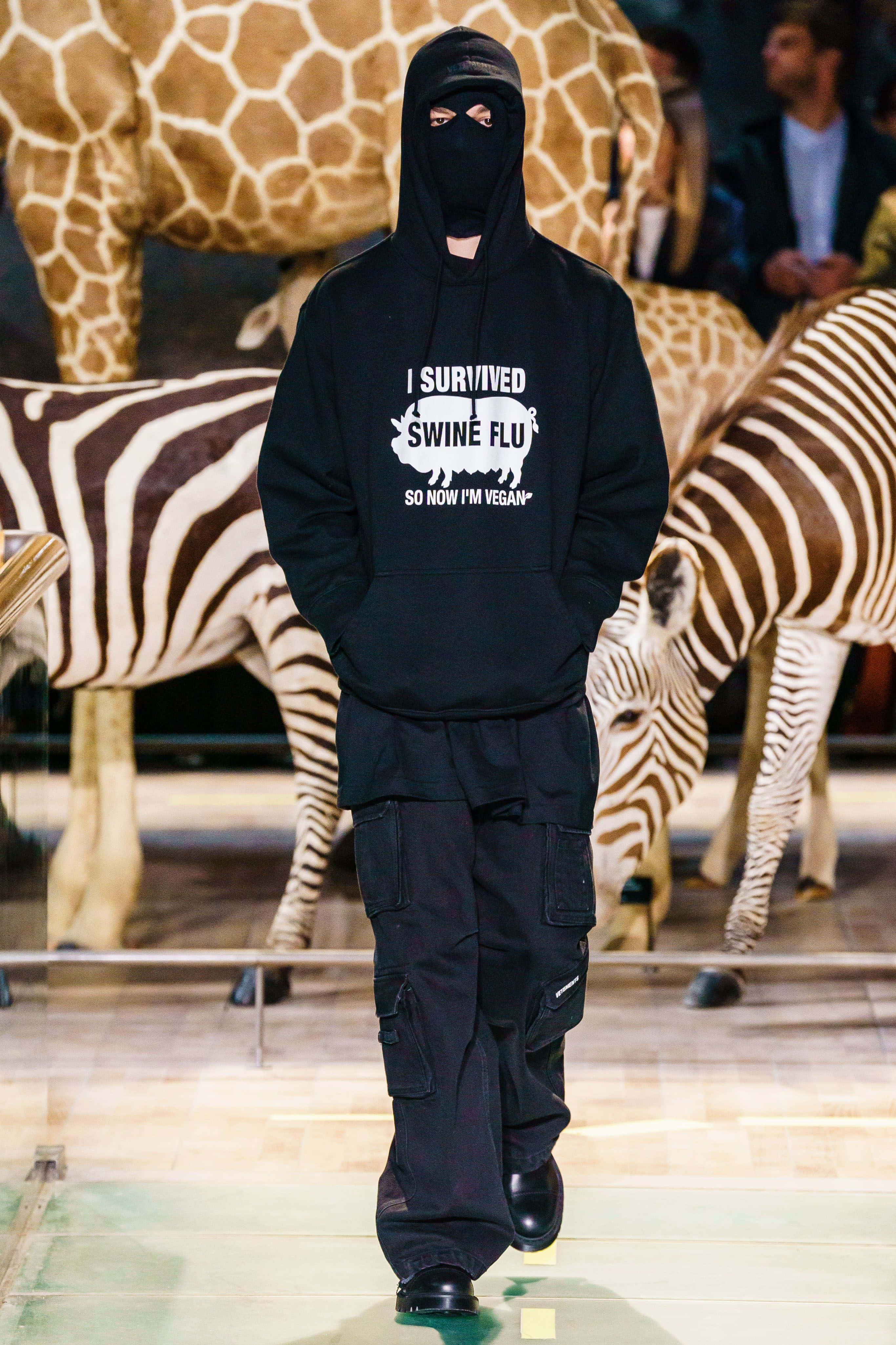 vetements 桌布 壁紙 — 一位模特兒穿著黑色連帽衫和工裝褲，在兩旁有真人大小的長頸鹿和斑馬雕像的走道上行走。