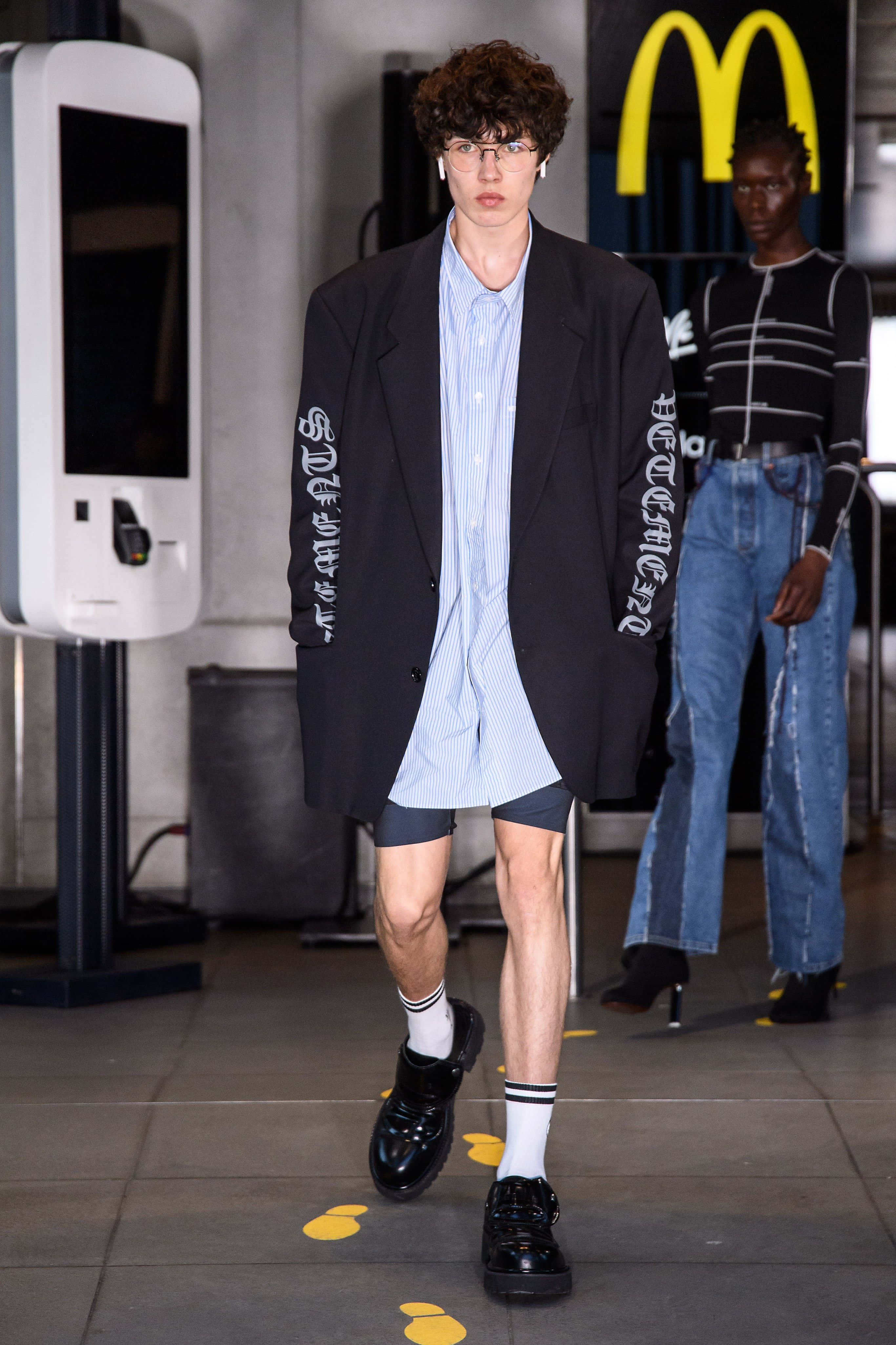vetements 桌布 壁紙 — 一位模特兒自信地穿著帶有白色字樣的黑色西裝外套，內搭條紋襯衫和短褲，在背景有麥當勞標誌的走秀台上行走。