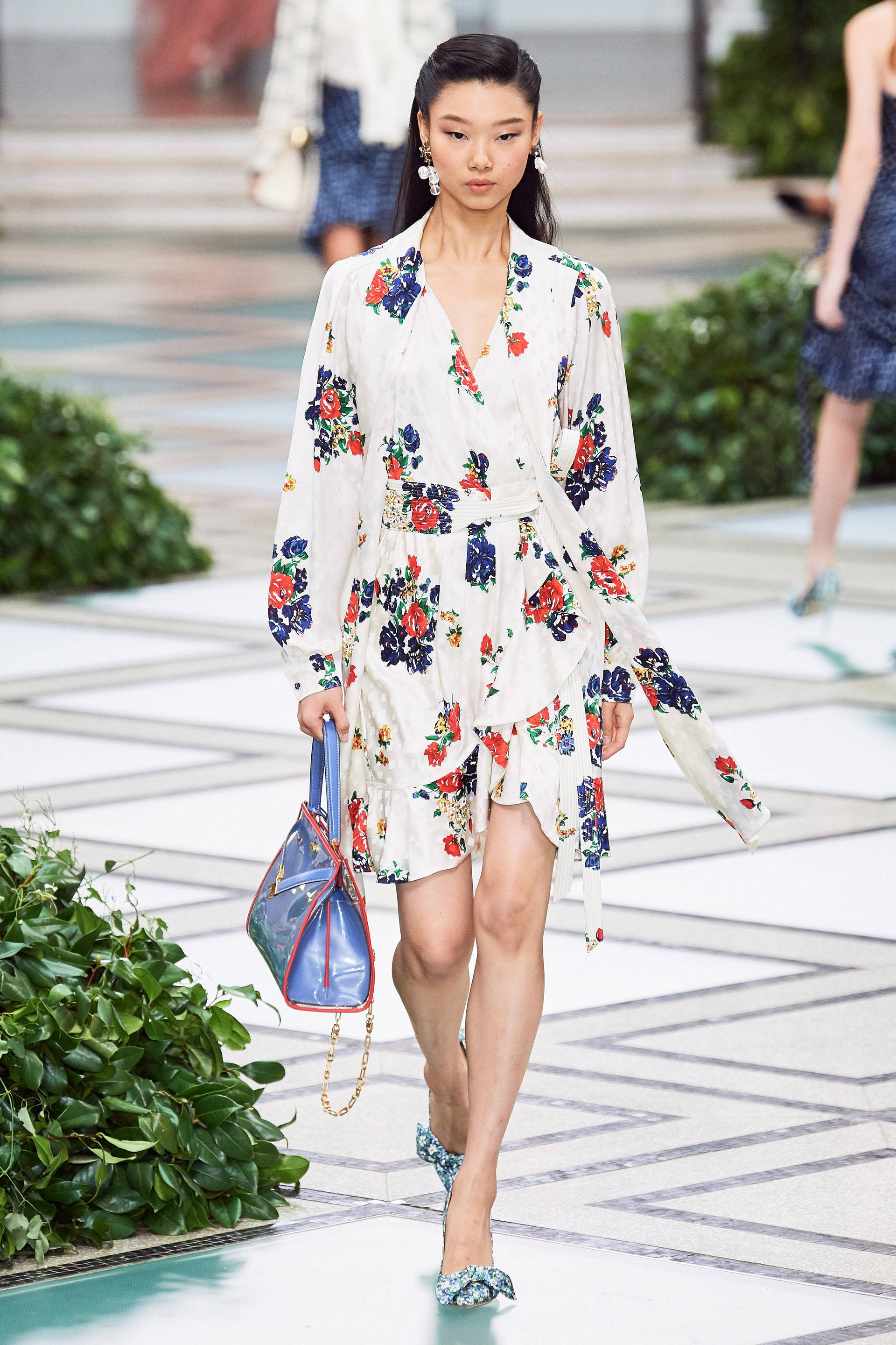 Tory Burch 桌布 壁紙 — 一位模特兒自信地穿著花卉印花裹身裙和圖案高跟鞋，在幾何瓷磚跑道兩旁綠植的襯托下走秀。