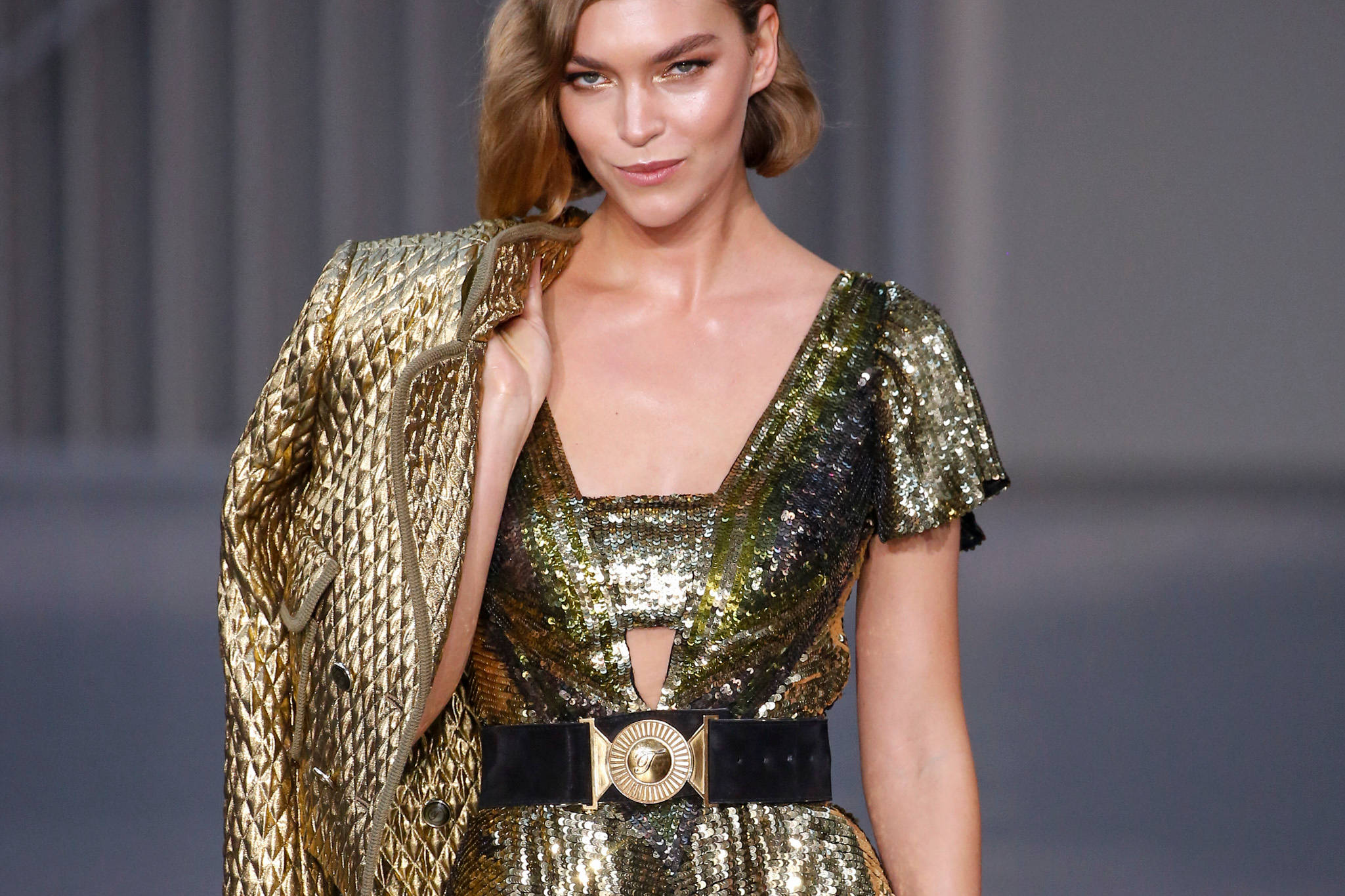Runway 桌布 壁紙 — 一位模特兒自信地穿著閃閃發光的金色亮片連衣裙和配套外套，搭配大膽的黑色腰帶，在時裝秀上走秀。