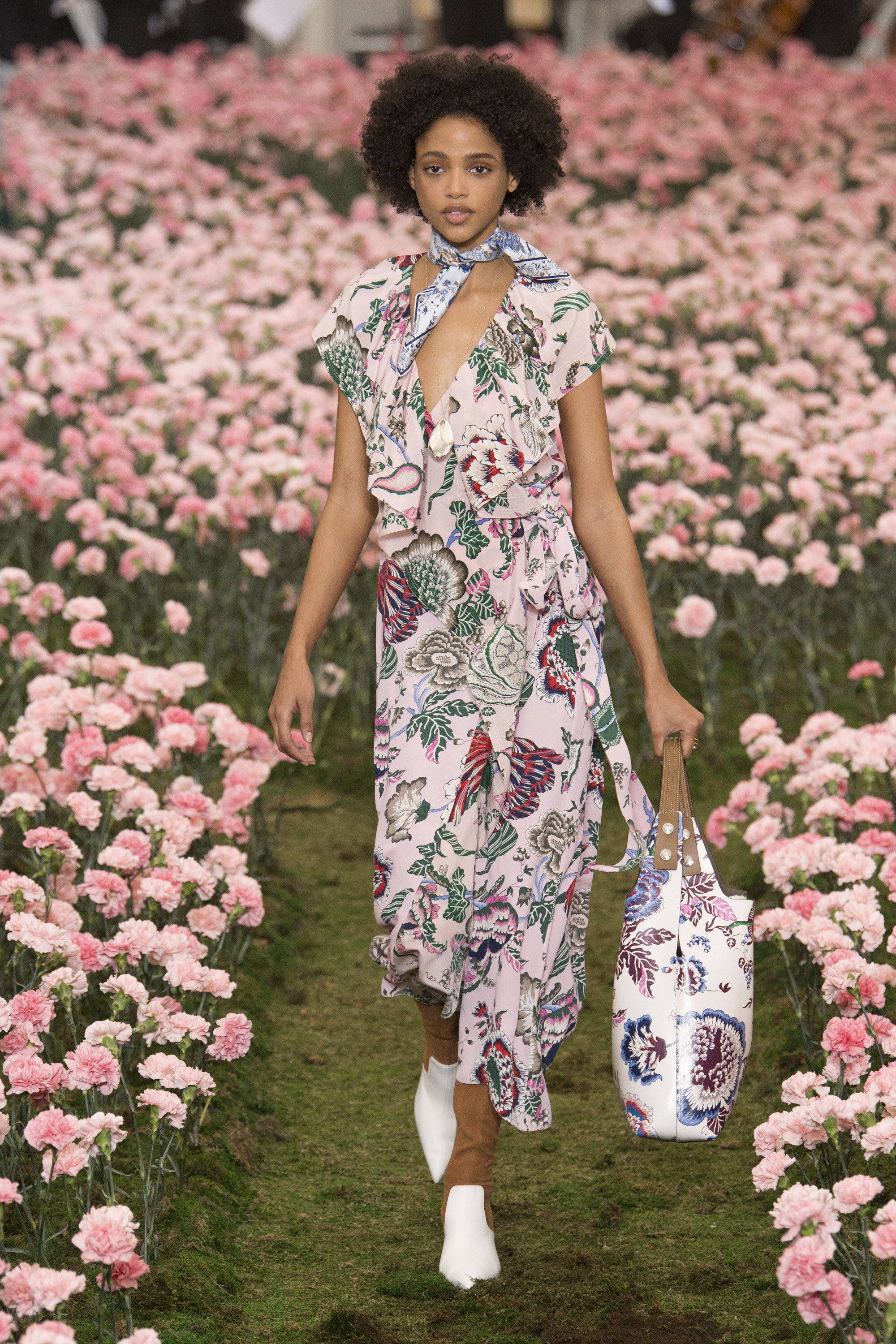 Tory Burch 桌布 壁紙 — 一位模特兒自信地穿過粉紅色康乃馨花田，身穿花卉洋裝並攜帶相配的手袋。