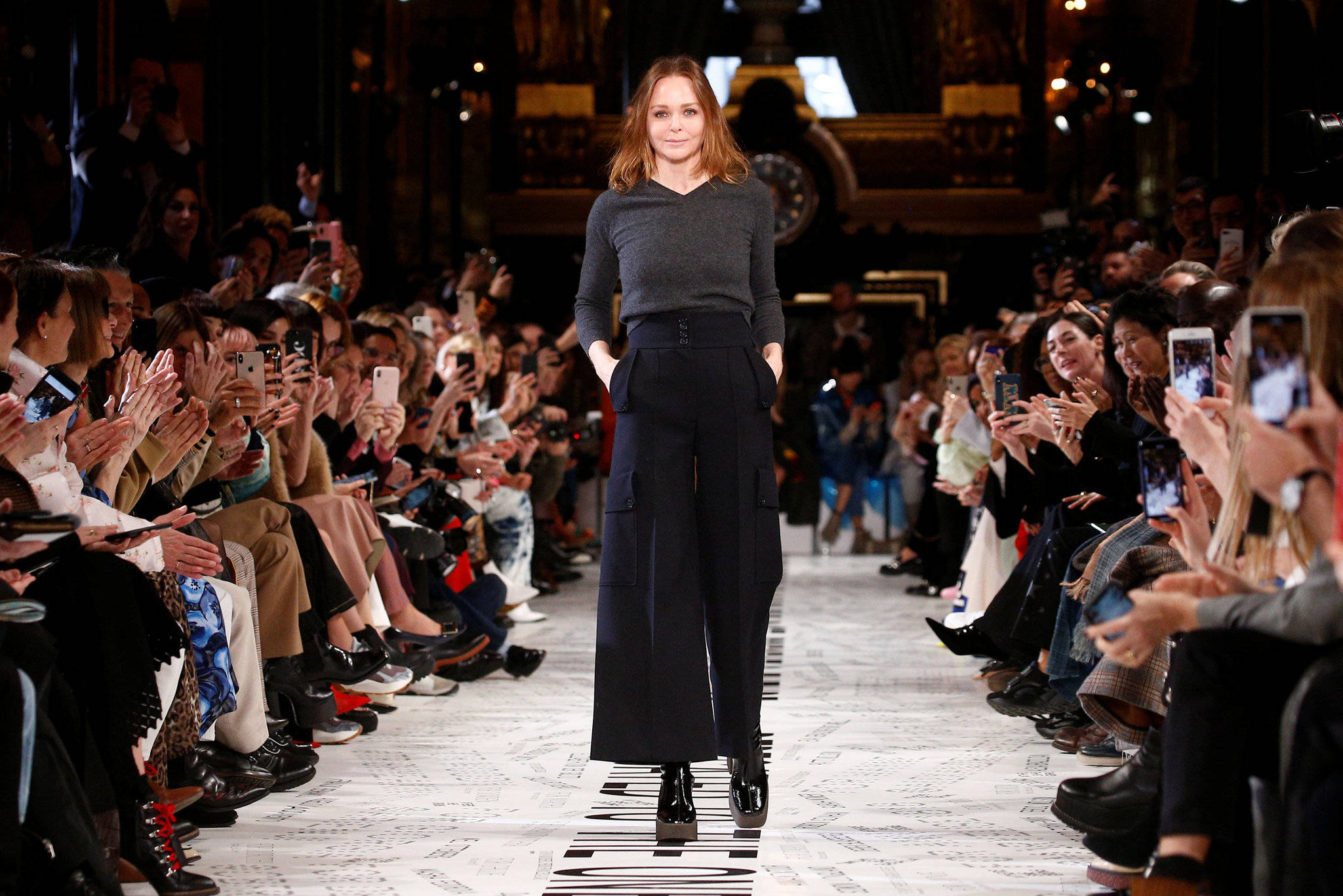Stella Mccartney 桌布 壁紙 — 一位模特兒自信地走在伸展台上，兩旁觀眾用手機捕捉這一刻。