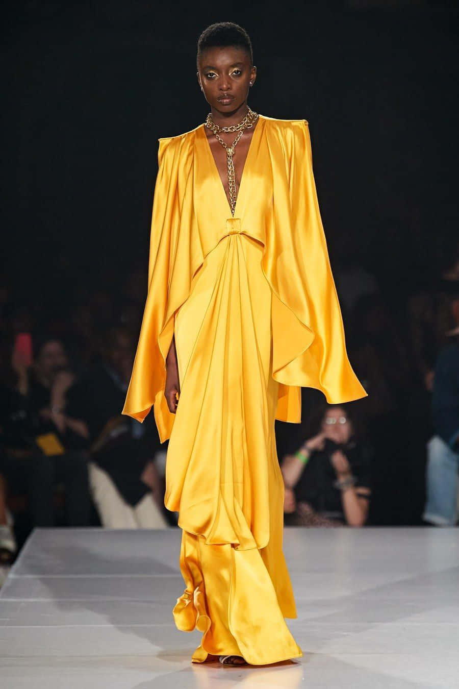 Spring Fashion 桌布 壁紙 — 一位模特兒自信地走在伸展台上，身穿搶眼的金黃色緞面長裙，搭配戲劇性的垂墜袖和深V領口。