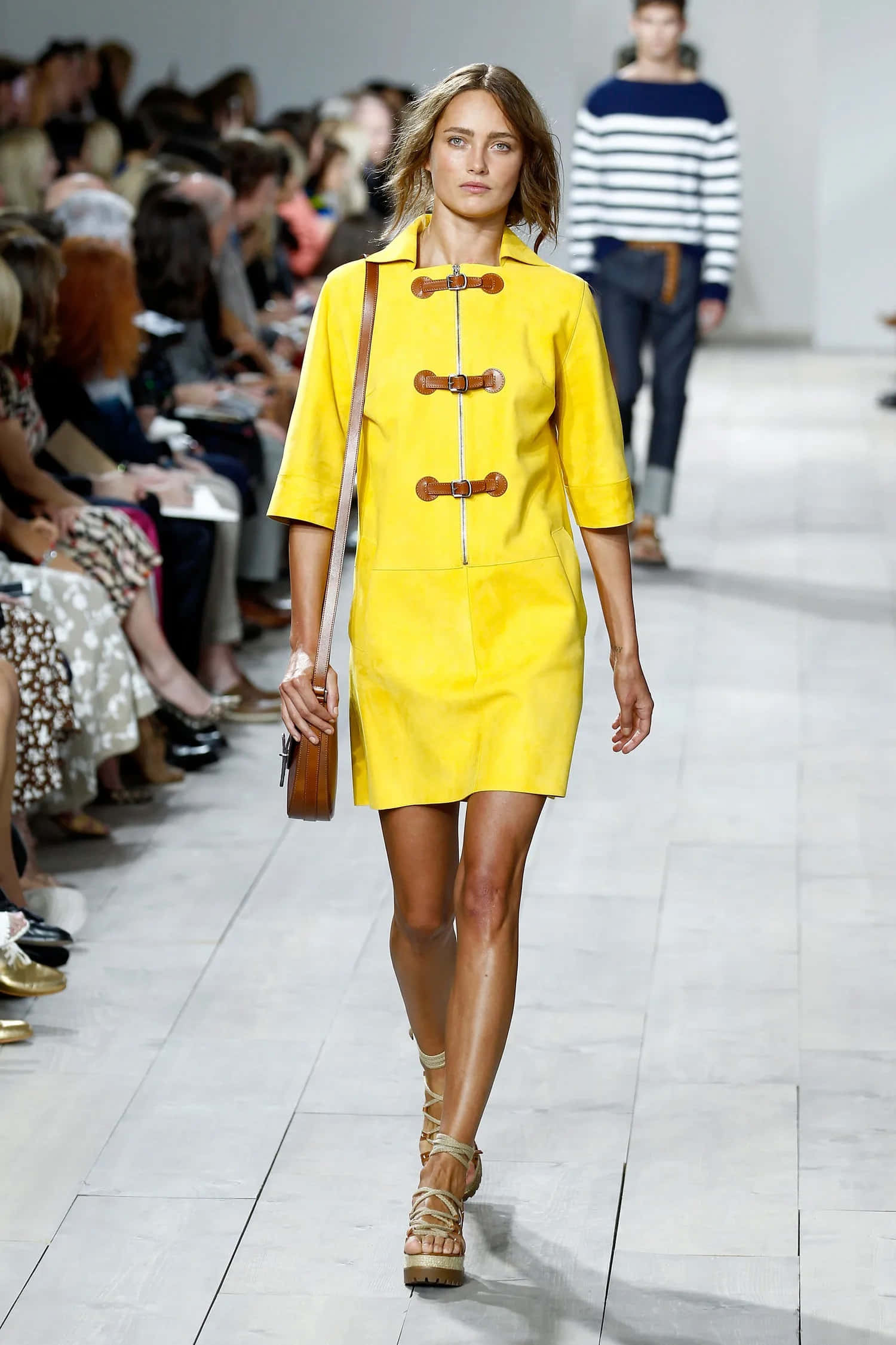 Yellow Fashion 桌布 壁紙 — 一位模特兒自信地走在伸展台上，身穿鮮黃色牛角扣大衣洋裝，搭配木質紐扣和棕褐色涼鞋。