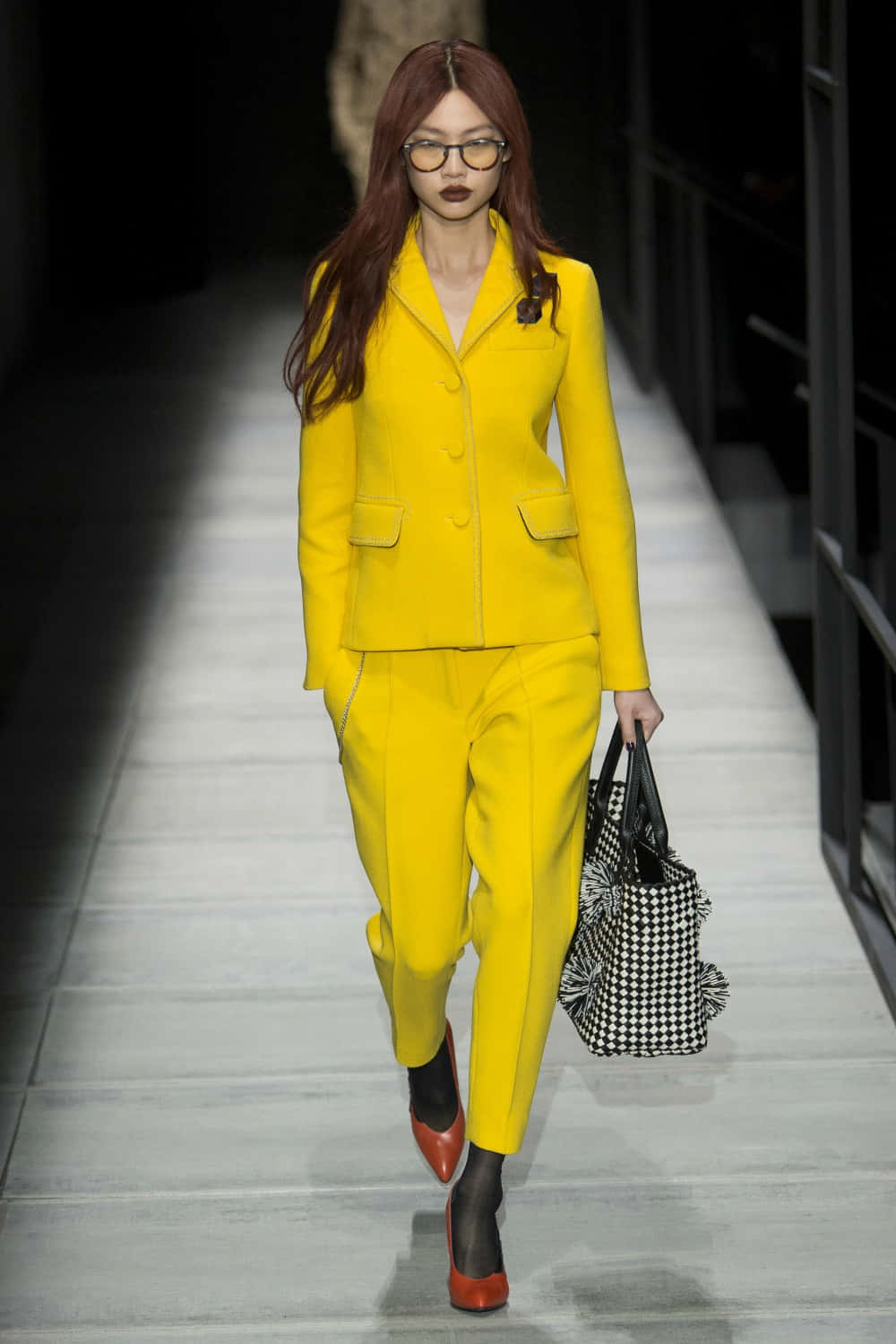 Yellow Fashion 桌布 壁紙 — 一位模特兒自信地走在伸展台上，身穿大膽的黃色西裝，手持黑白格紋包包。