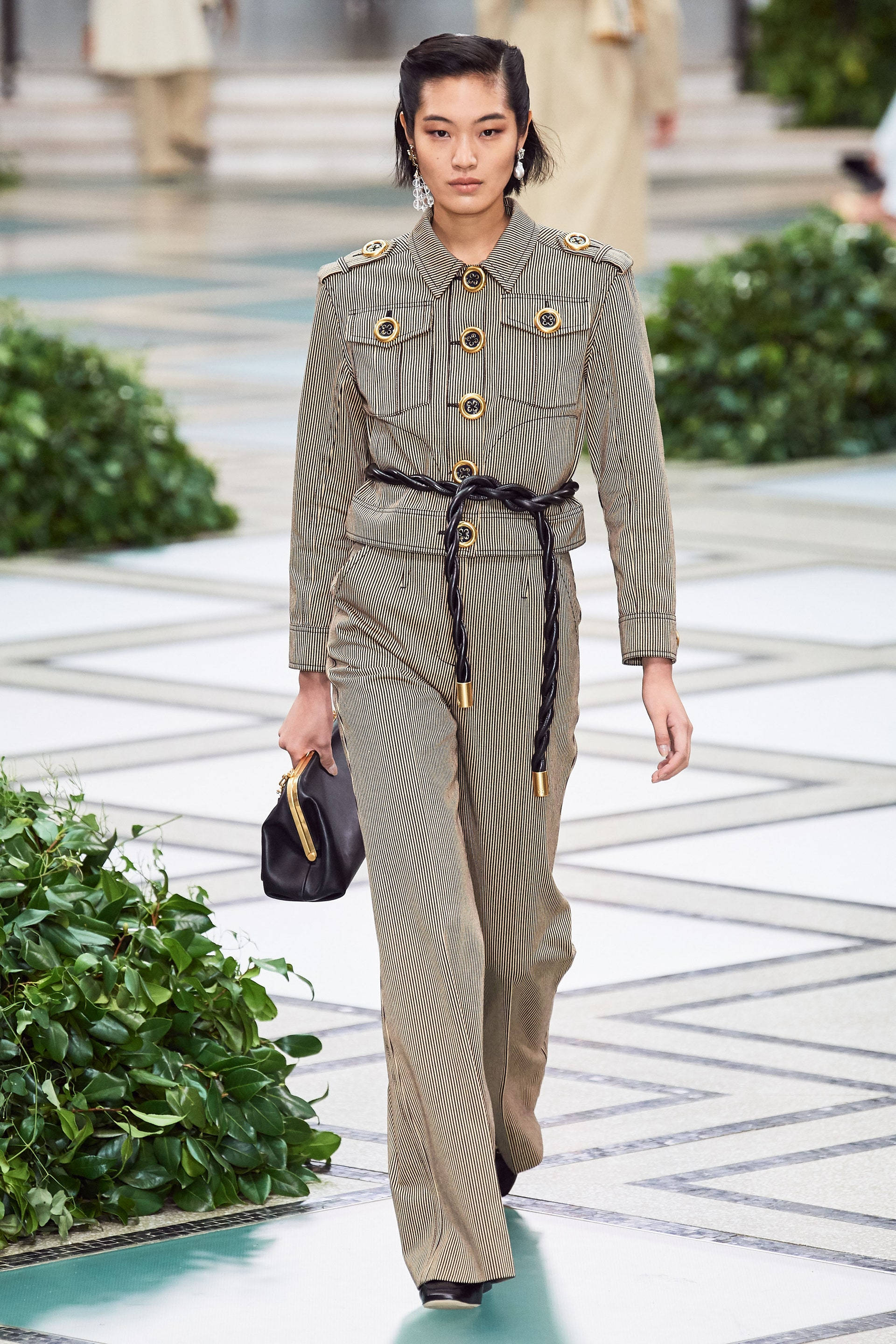 Tory Burch 桌布 壁紙 — 一位模特兒自信地走在伸展台上，身穿帶有金色鈕扣和編織腰帶的細條紋軍裝連身褲。