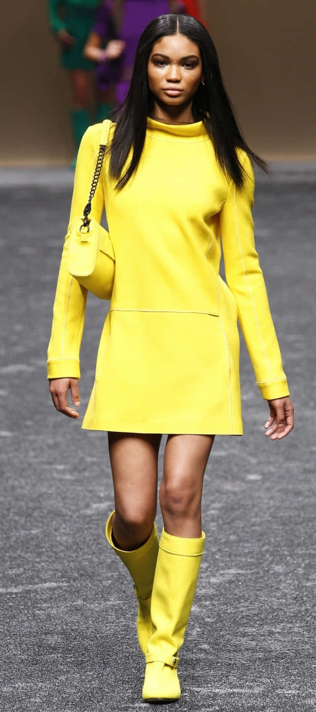 Yellow Fashion 桌布 壁紙 — 一位模特兒自信地走在伸展台上，身穿大膽的黃色套裝，包括合身連身裙、及大腿的靴子和相配的手提包。