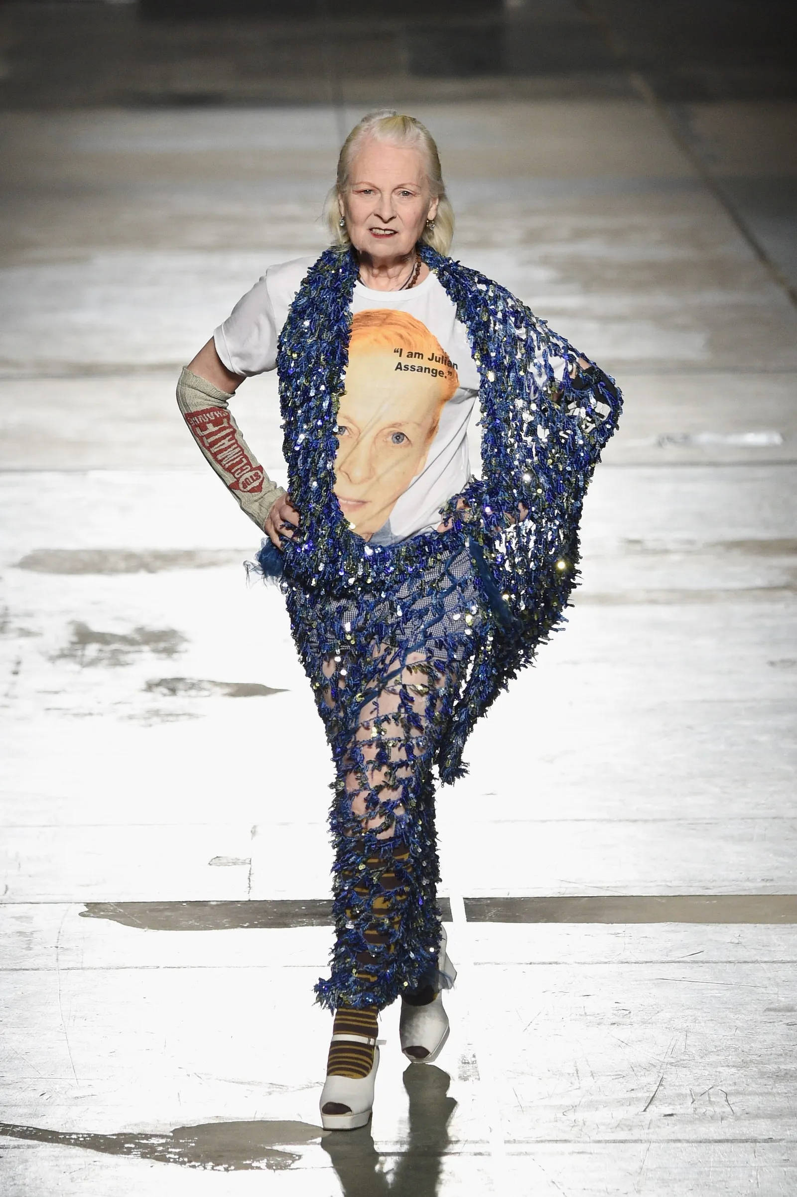 Vivienne Westwood 桌布 壁紙 — 一位模特兒自信地走在伸展台上，身穿印有臉孔的白色T恤和閃亮的藍色亮片套裝。