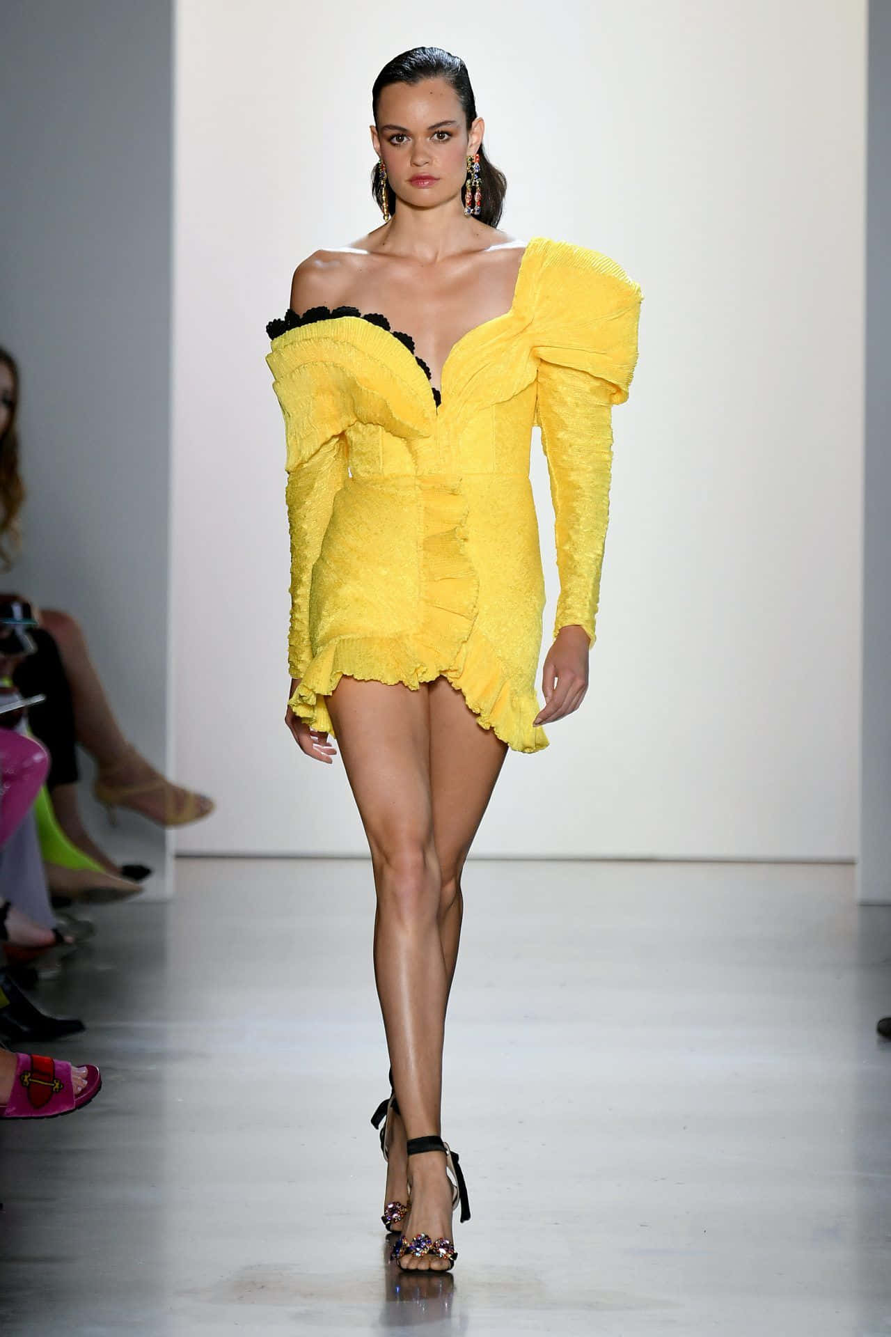 Yellow Fashion 桌布 壁紙 — 一位模特兒自信地走在伸展台上，身穿大膽的黃色荷葉邊迷你裙，搭配黑色飾邊和高跟鞋。