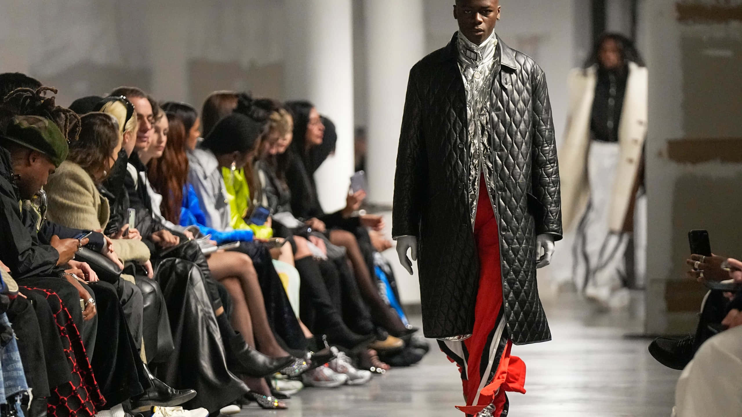 Runway 桌布 壁紙 — 一位模特兒自信地走在伸展台上，身穿黑色絎縫外套，內搭紅白套裝，坐著的觀眾正在觀看。