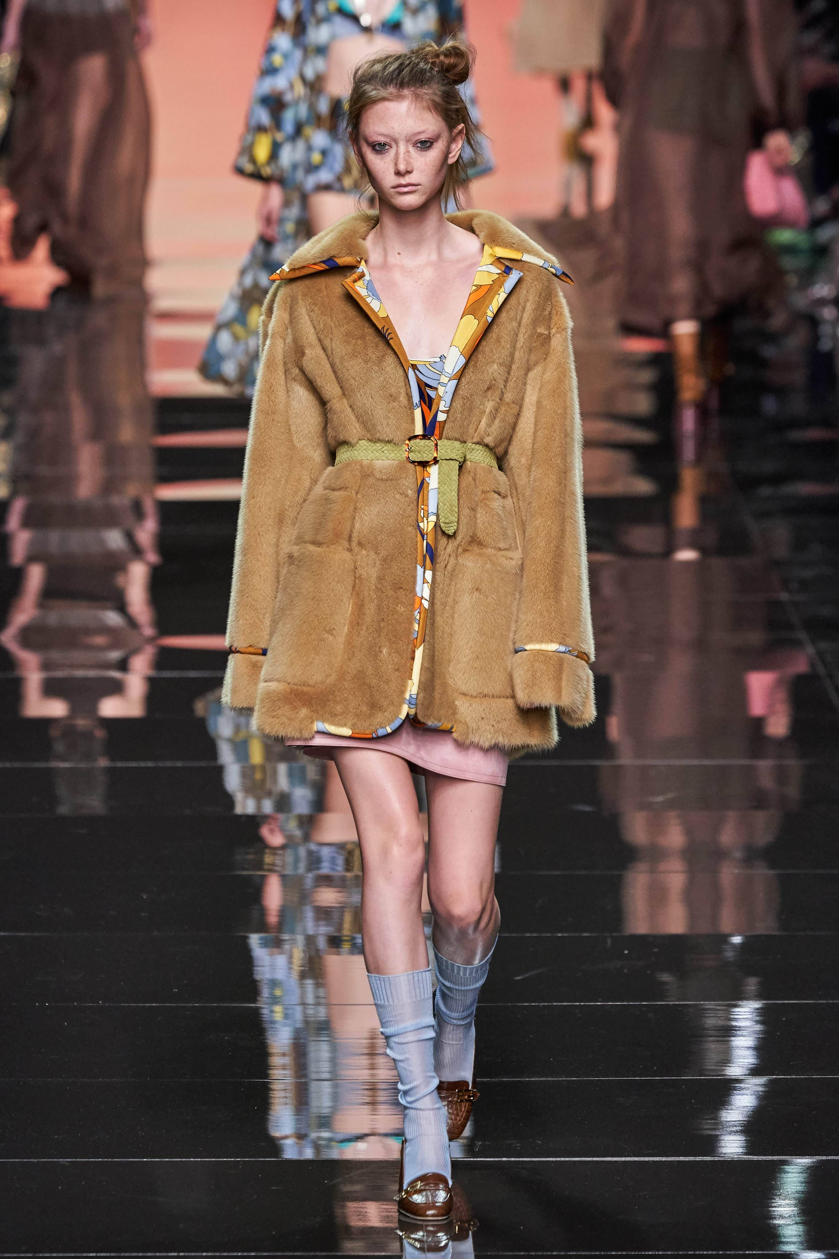 Fendi 桌布 壁紙 — 一位模特兒自信地走在光亮的伸展台上，身穿帶有圖案飾邊的棕褐色毛皮大衣和淺藍色襪子。