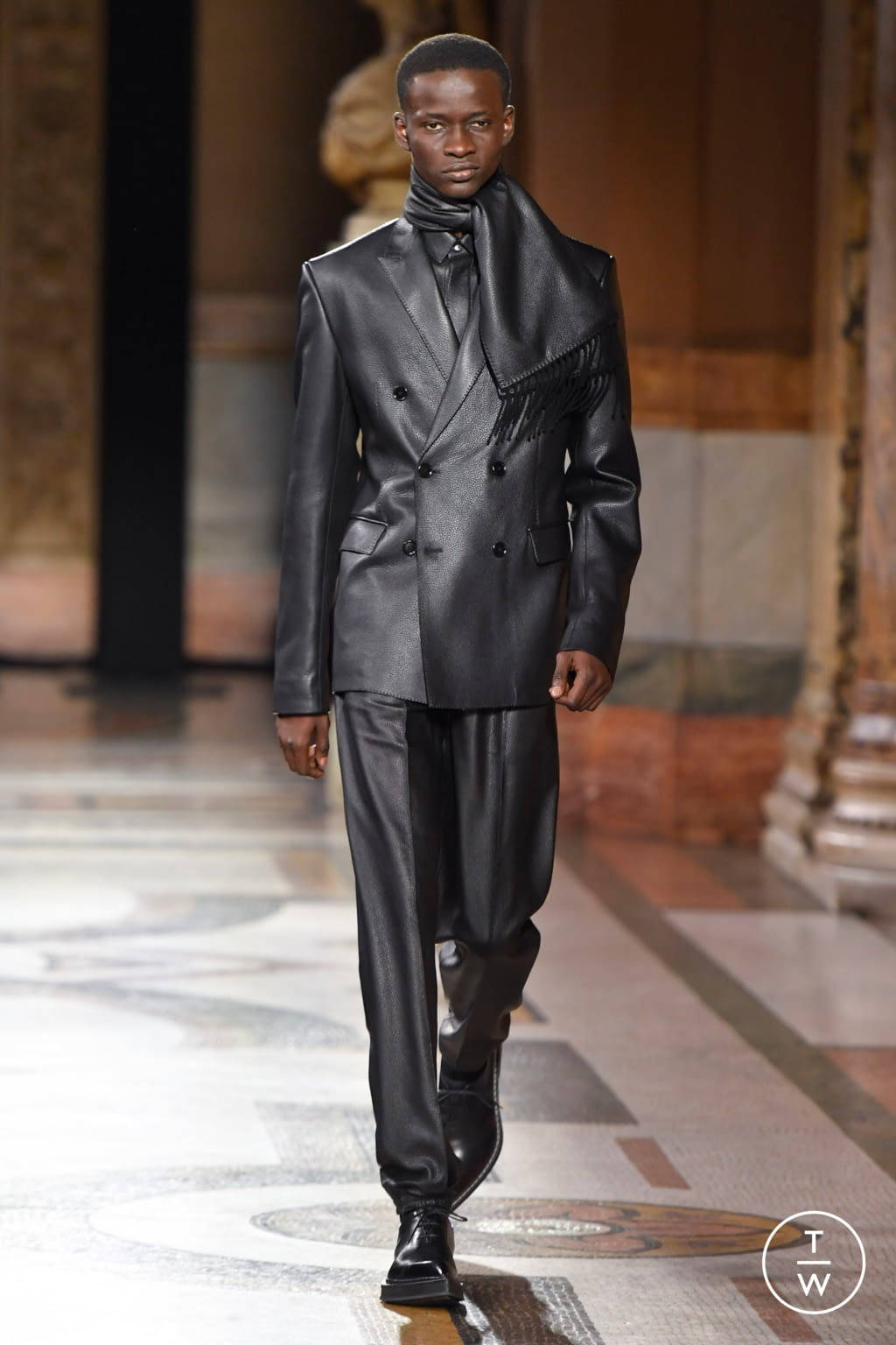 Runway 桌布 壁紙 — 一位模特兒自信地走在大理石地板的時裝伸展台上，身穿時尚黑色皮革雙排扣西裝，帶有流蘇細節。