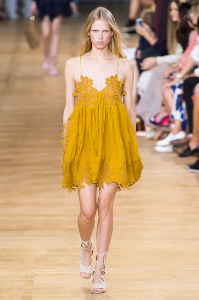 Yellow Fashion 桌布 壁紙 — 一位模特兒自信地走在木製伸展台上，身穿芥末黃色蕾絲連衣裙和白色繫帶高跟鞋。