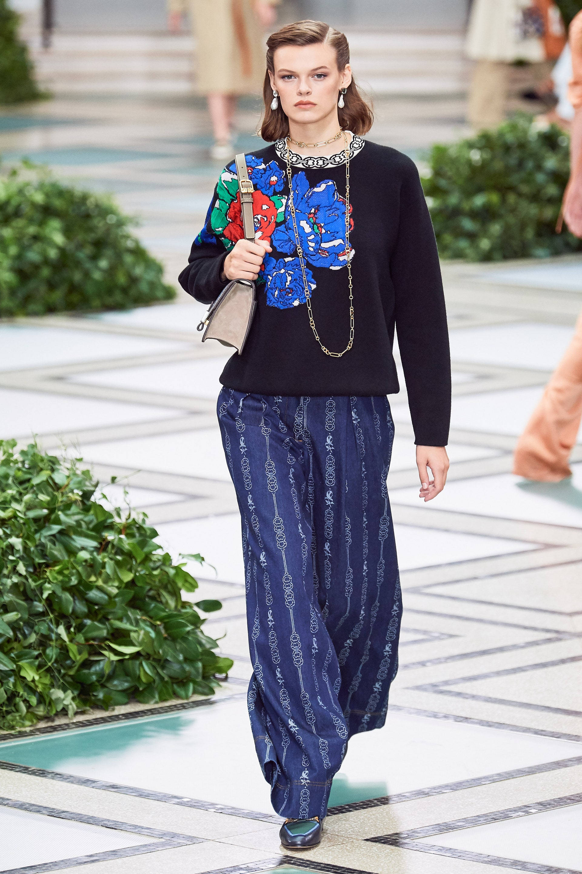 Tory Burch 桌布 壁紙 — 一位模特兒自信地走在瓷磚跑道上，兩旁是綠色樹籬，身穿黑色毛衣，帶有鮮明的藍色花卉刺繡和圖案藍色褲子。
