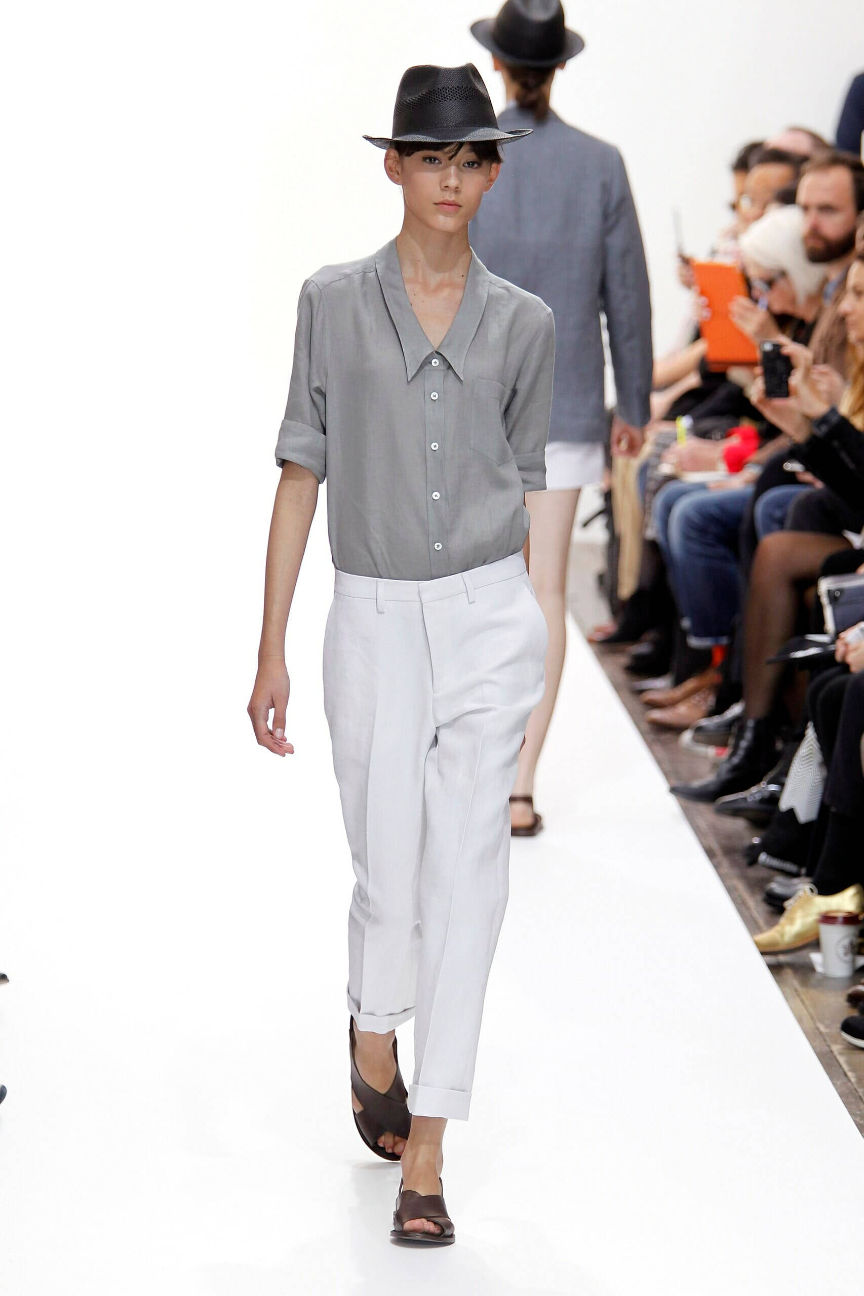Fashion Model 桌布 壁紙 — 一位模特兒自信地走在白色伸展台上，身穿灰色襯衫、白色褲子和黑色帽子，觀眾從兩側觀看。