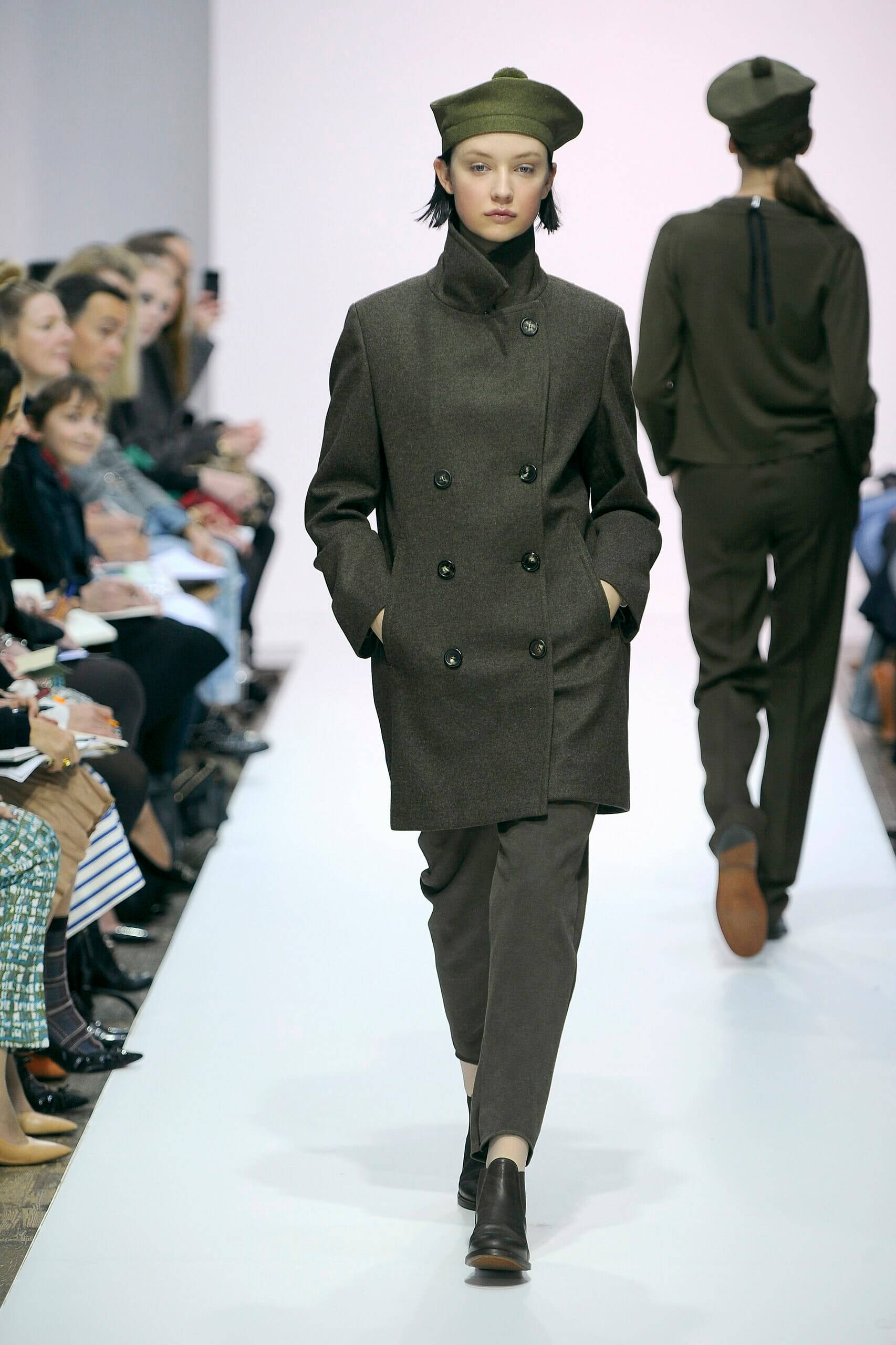Fashion Model 桌布 壁紙 — 一位模特兒自信地走在白色伸展台上，身穿軍裝風格的橄欖綠色外套和貝雷帽，觀眾在旁觀看。