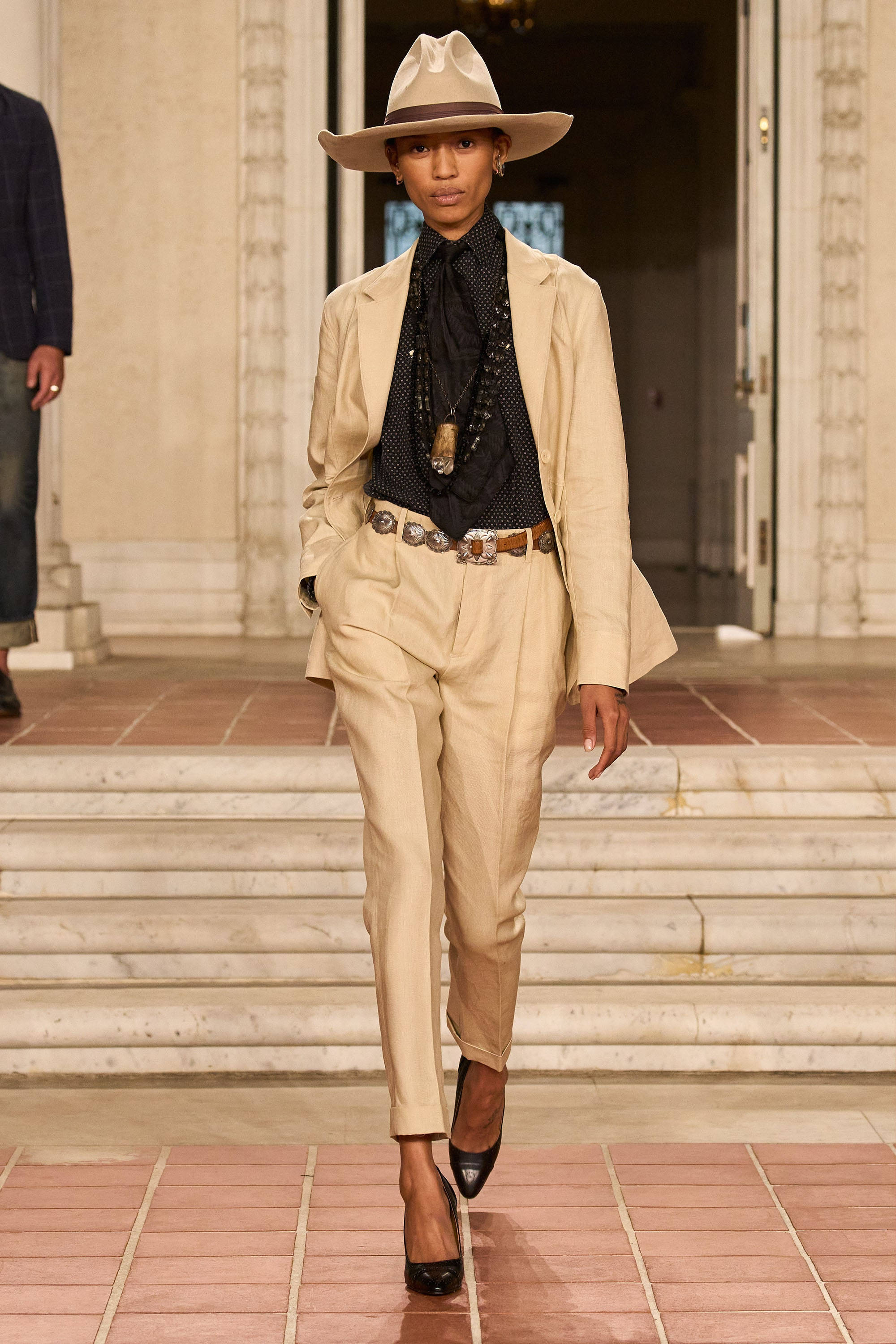 ralph lauren 桌布 壁紙 — 一位模特兒自信地走在磁磚走秀台上，身穿米色西裝、牛仔帽和黑色高跟鞋。