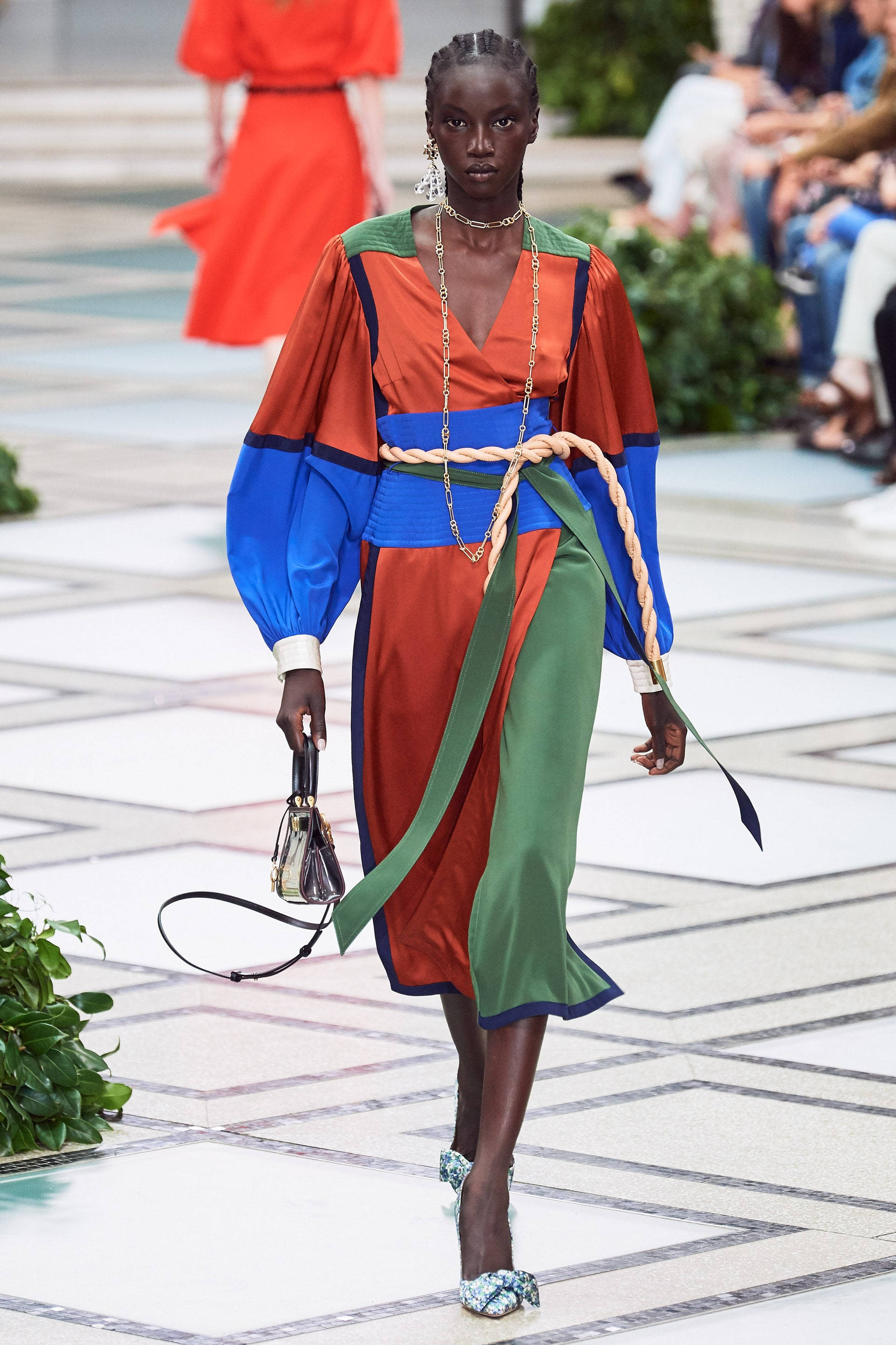 Tory Burch 桌布 壁紙 — 一位模特兒自信地走在磁磚走秀台上，身穿大膽的橙色、藍色和綠色拼色連衣裙，搭配編織繩腰帶和圖案高跟鞋。