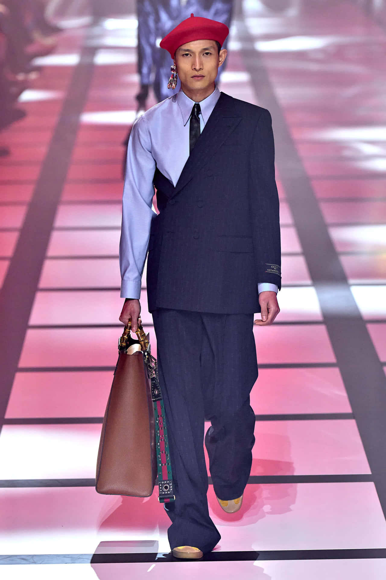 Gucci 桌布 壁紙 — 一位模特兒自信地走在粉紅色和黑色瓷磚的T台上，身穿海軍藍西裝、紅色貝雷帽，並攜帶一個棕色皮革手提包。