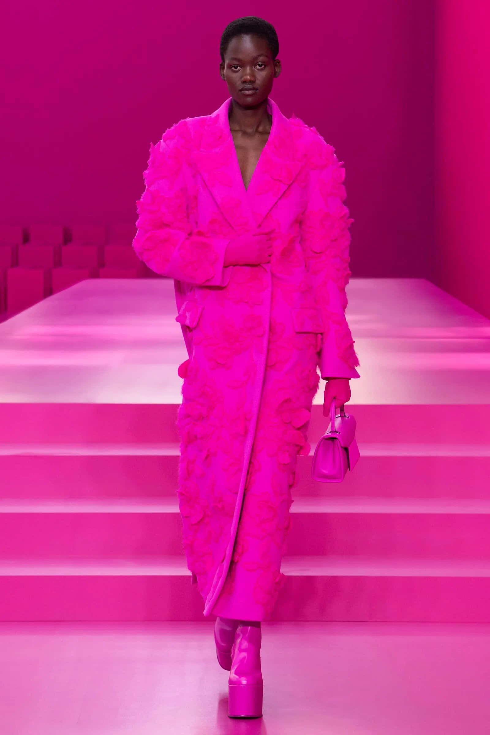 Pink Fashion 桌布 壁紙 — 一位模特兒自信地走在粉紅色的伸展台上，身穿一件寬鬆的粉紅色花卉外套和相配的配件。