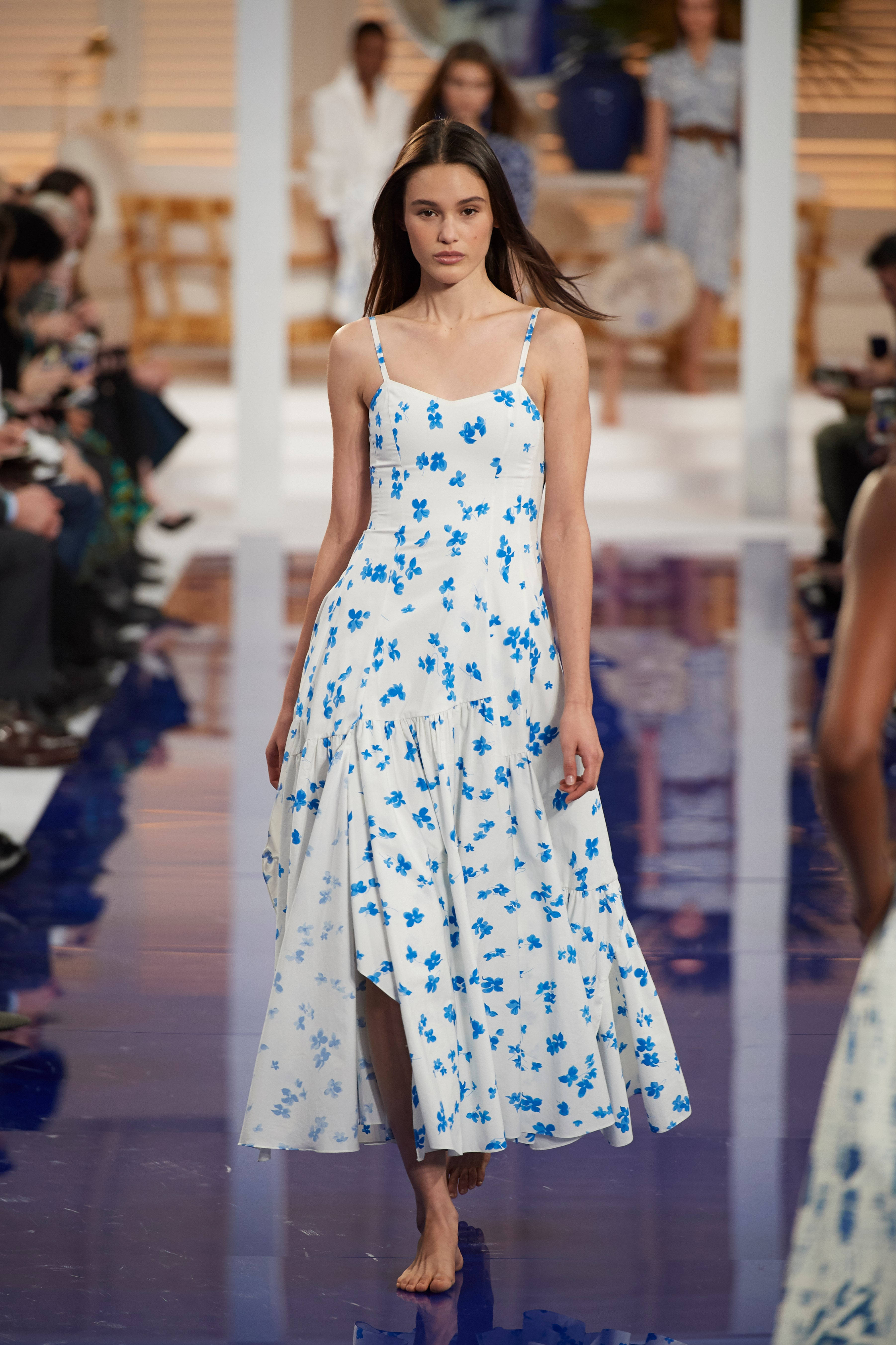 Runway 桌布 壁紙 — 一位模特兒自信地走在閃亮的伸展台上，身穿白色花卉洋裝，點綴藍色花朵，在明亮的時裝秀燈光下。