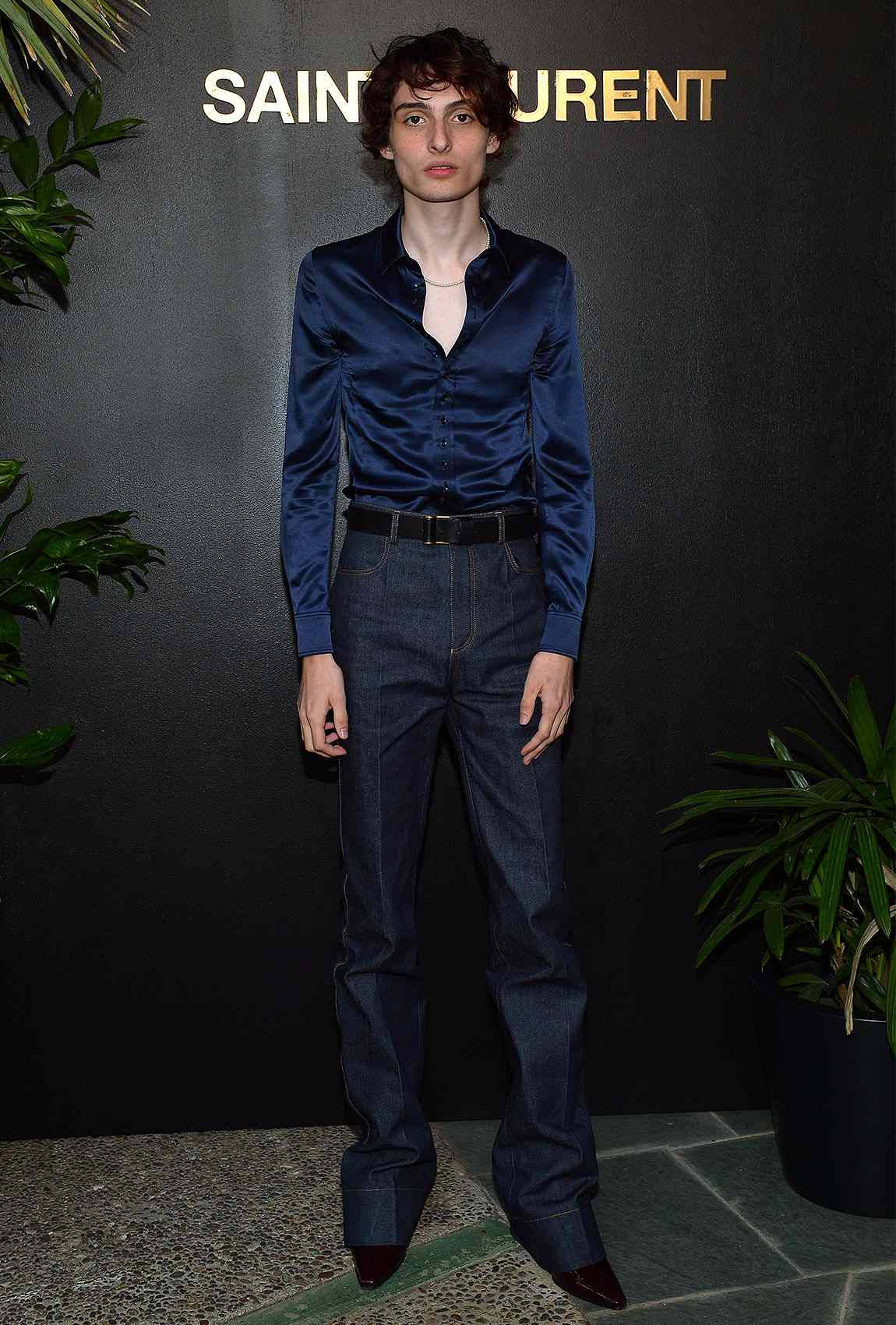 finn wolfhard 桌布 壁紙 — 一位模特兒身穿海軍藍緞面襯衫和喇叭牛仔褲，在帶有'SAINT LAURENT'標誌和盆栽植物的深色牆壁前擺姿勢。