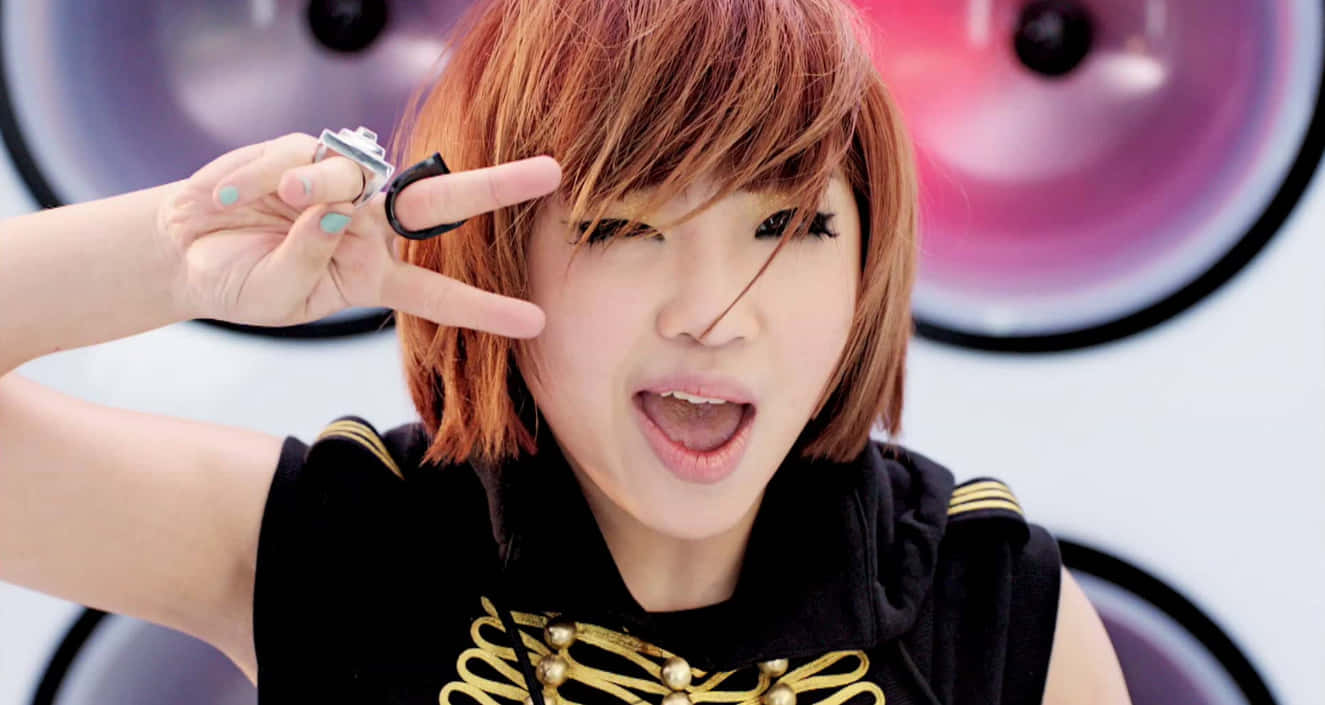 2NE1 桌布 壁紙 — 一位橘髮年輕女性在巨型卡通眼睛背景下眨眼並做出和平手勢。