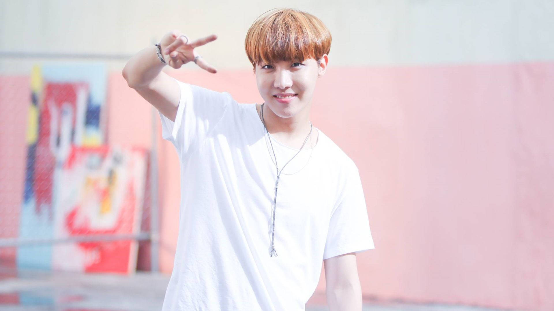 jhope 桌布 壁紙 — 一位橘髮年輕男子在色彩繽紛的塗鴉牆前微笑並比出和平手勢。