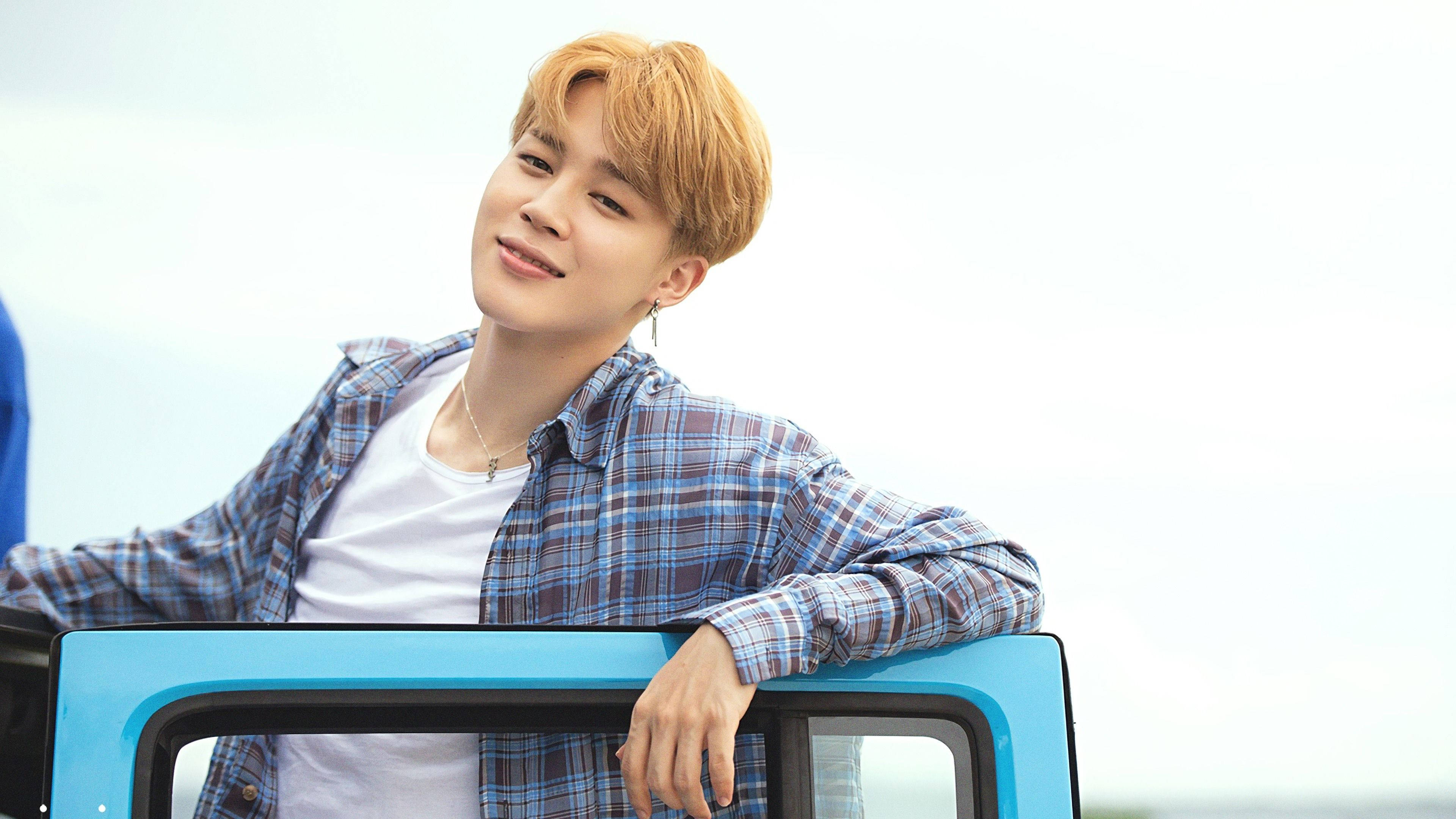 Jimin Bts Cute 桌布 壁紙 — 一位橘髮年輕男子隨意靠在一輛亮藍色車門上，在蒼白的天空下溫柔微笑。
